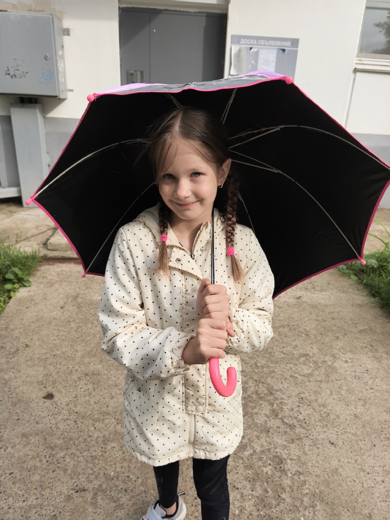 A young girl stands outdoors, holding a black umbrella with a pink trim and handle. She wears a white jacket with black polka dots, black leggings, and white sneakers. Her hair is styled in braids secured with pink ties. The background includes a concrete surface, a building with a grey door, and a notice board with Russian text. This is in the city of Chaikovsky, Russia.