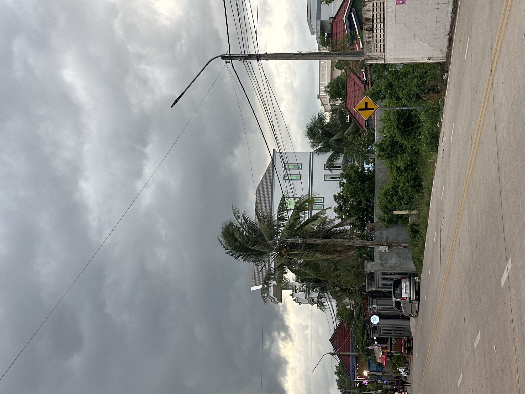In Kampot, Cambodia, a cloudy day creates a somber atmosphere over a street scene. A gray sky, heavy with impending rain, looms above. A white multi-story building with green-tinted windows stands prominently, flanked by lush greenery and palm trees. A streetlight with numerous wires stretches across the frame.
On the street, a silver hatchback car is parked near a gated entrance, its tires slightly turned. A tuk-tuk is visible further down the road, and a few people walk along the sidewalk. A yellow diamond-shaped traffic sign with a black T-intersection symbol is mounted on a pole. The road is paved and marked with yellow and white lines. Buildings with red roofs are visible in the background, along with dense vegetation. The scene appears to be a typical urban street with a mix of residential and commercial properties, under a dramatic, overcast sky.
