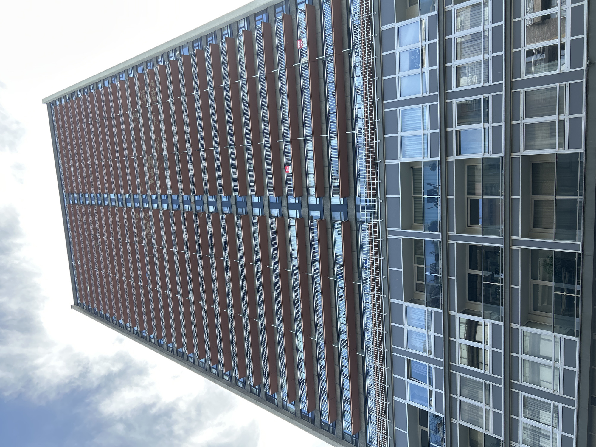 A low-angle, full-frame exterior shot captures a towering, modern apartment building against a cloudy sky in Ostend, Belgium. The building is characterized by repetitive horizontal balconies with white railings and terracotta-colored soffits, punctuated by numerous rectangular windows. The lower floors feature a different architectural style, with larger windows arranged in grid patterns and some recessed balconies with glass railings. The overall impression is one of a dense urban environment with a focus on residential architecture.