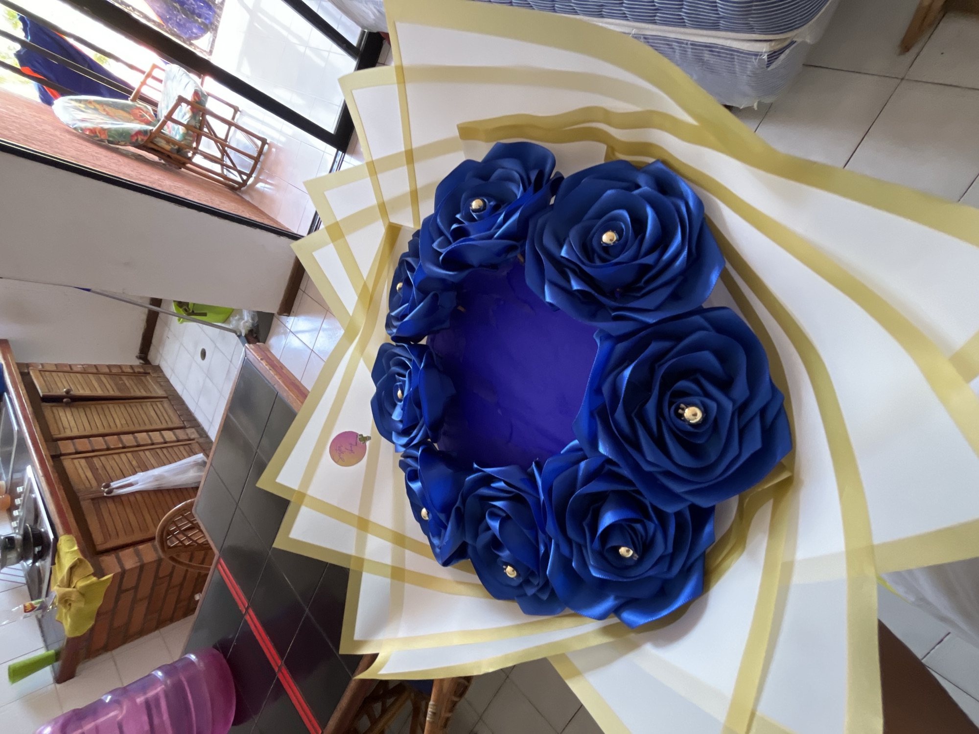 This image captures a vibrant, circular arrangement of artificial royal blue roses, presented in a bouquet-like fashion, set within a residential interior in Porlamar, Venezuela. The roses, crafted from a lustrous, satin-like fabric, each feature a small, golden bead at their center, adding a touch of elegance. They are encircled by white wrapping paper or cardstock accented with wide, gold stripes or ribbons, fanning outwards from the central rose display. The center of the rose arrangement itself is a deep blue, circular void.
The photograph is taken from a slightly high angle, looking down, causing the background elements to appear tilted. To the left, various domestic items are visible, including wooden cabinets or counters, a stainless steel sink with a yellow cloth, and a large purple water bottle on a tiled floor. Further back, a metal railing and a wooden chair with a colorful, patterned cushion are seen, suggesting a window or balcony view. To the upper right, part of what appears to be a mattress or striped fabric object is visible, along with more tiled flooring. Bright natural light illuminates the scene, indicating it is daytime. A small, circular tag attached to the rose arrangement is partially visible, with the text "Love with..." discernable.