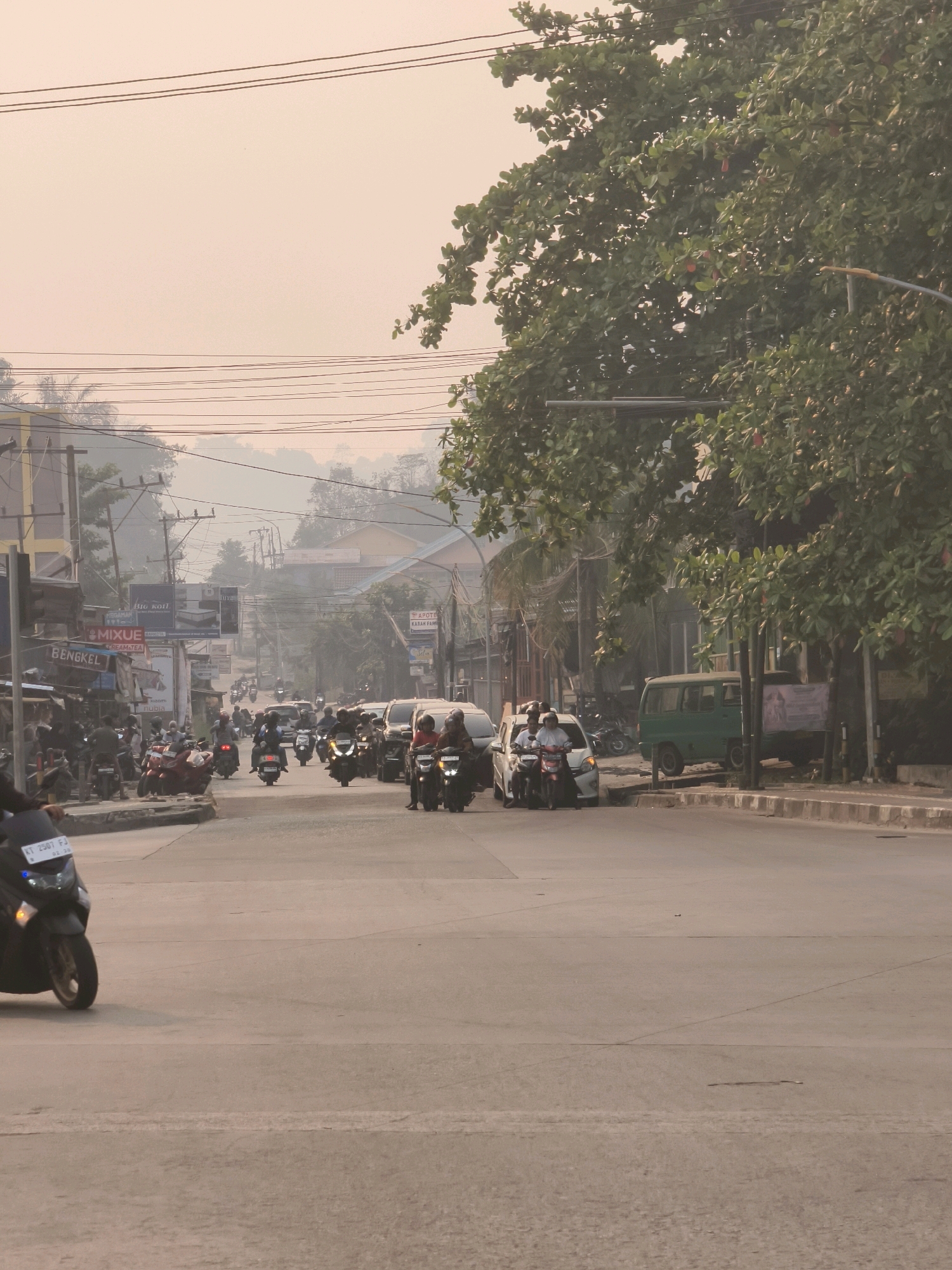 The image captures a street scene in Samarinda, Indonesia, under a hazy sky, likely due to pollution or a natural phenomenon. The foreground is dominated by a paved road with traffic markings. Several motorcycles are present, with one prominently in the lower left corner, a dark-colored scooter with a license plate reading "KT 2507 PJ". In the middle ground, a procession of motorcycles and a few cars navigate the road. On the left side of the street, buildings and businesses are visible, including signs for "BENGKEL" (workshop) and "MIXUE ICE CREAM TEA". On the right side, a green van is parked, and more trees line the roadside. Power lines crisscross the sky above the street. The overall atmosphere suggests a busy urban environment during daylight hours, with a muted color palette due to the haze.