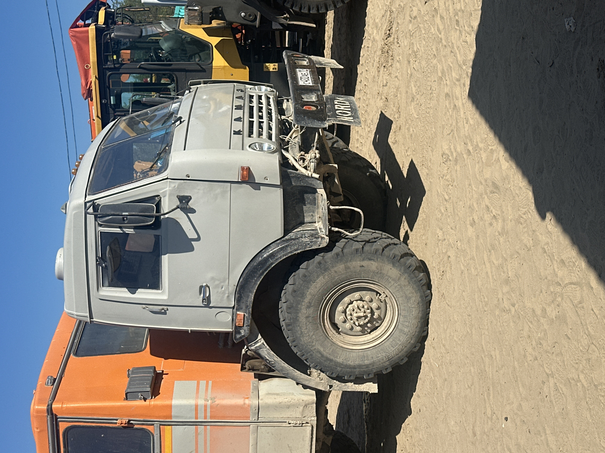 The image shows two vehicles parked on a sandy surface under a clear blue sky. The foreground is dominated by the front portion of a gray truck with an orange truck body behind it. The gray truck has large, rugged tires, a prominent front grille with the word "KAMAZ" visible, and a black license plate with the characters "C432AE 11". Below the license plate, another black plate reads "NORD". A portion of a yellow construction vehicle with a red canopy is visible to the right of the gray truck. The environment appears to be an outdoor area, possibly a construction site or a storage yard, with sand covering the ground and tire tracks visible. No people are clearly depicted in the scene, though a reflection in the gray truck's window suggests the presence of at least one person inside. The lighting suggests it is daytime, with strong sunlight casting shadows. The location is identified as Chikshino, Russia.