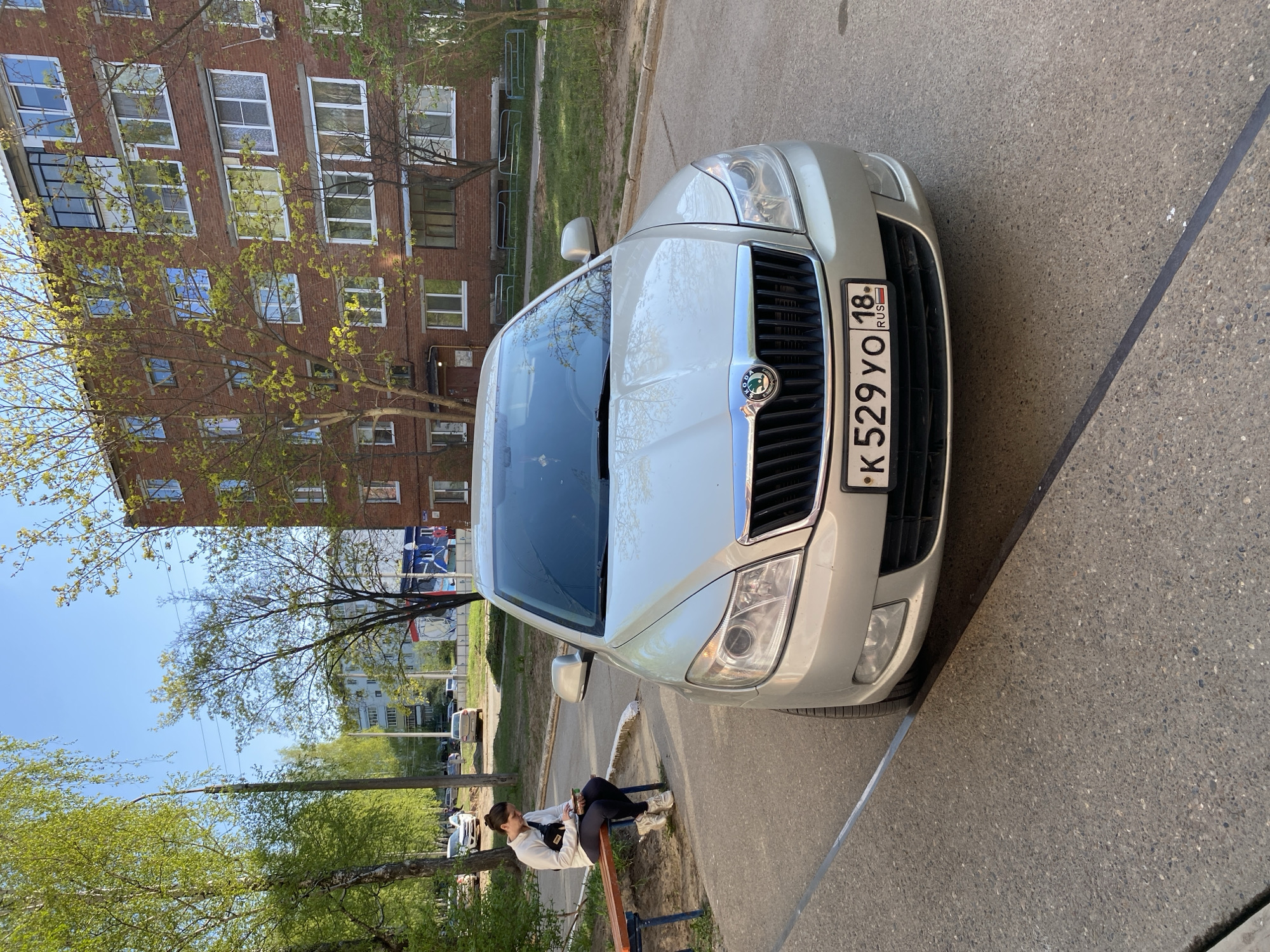 A silver Skoda car is parked on a paved surface next to a curb. A person is sitting on a park bench near the car. In the background, there is a multi-story red brick building, trees with green foliage, and other buildings in the distance. The car's license plate is visible, indicating it is from Russia, specifically region 18. The sign on the license plate gives the impression the location is possibly the city of Noviy, Russia.