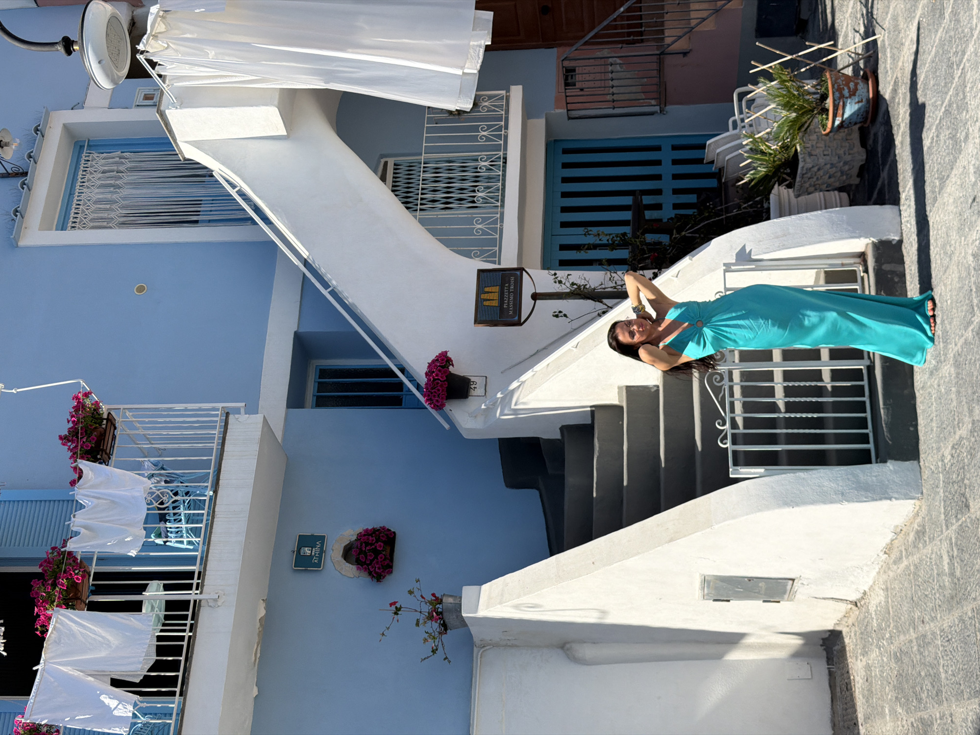 The image shows a woman in a turquoise dress standing on a white concrete staircase in Procida, Italy. The architecture features white and blue buildings with flower boxes. Clothes hang drying on a balcony in the background. There are two wall-mounted signs, one with the name "Ricetta di Massimo Troisi" and another with "Athina." The environment is a sunlit, narrow street.