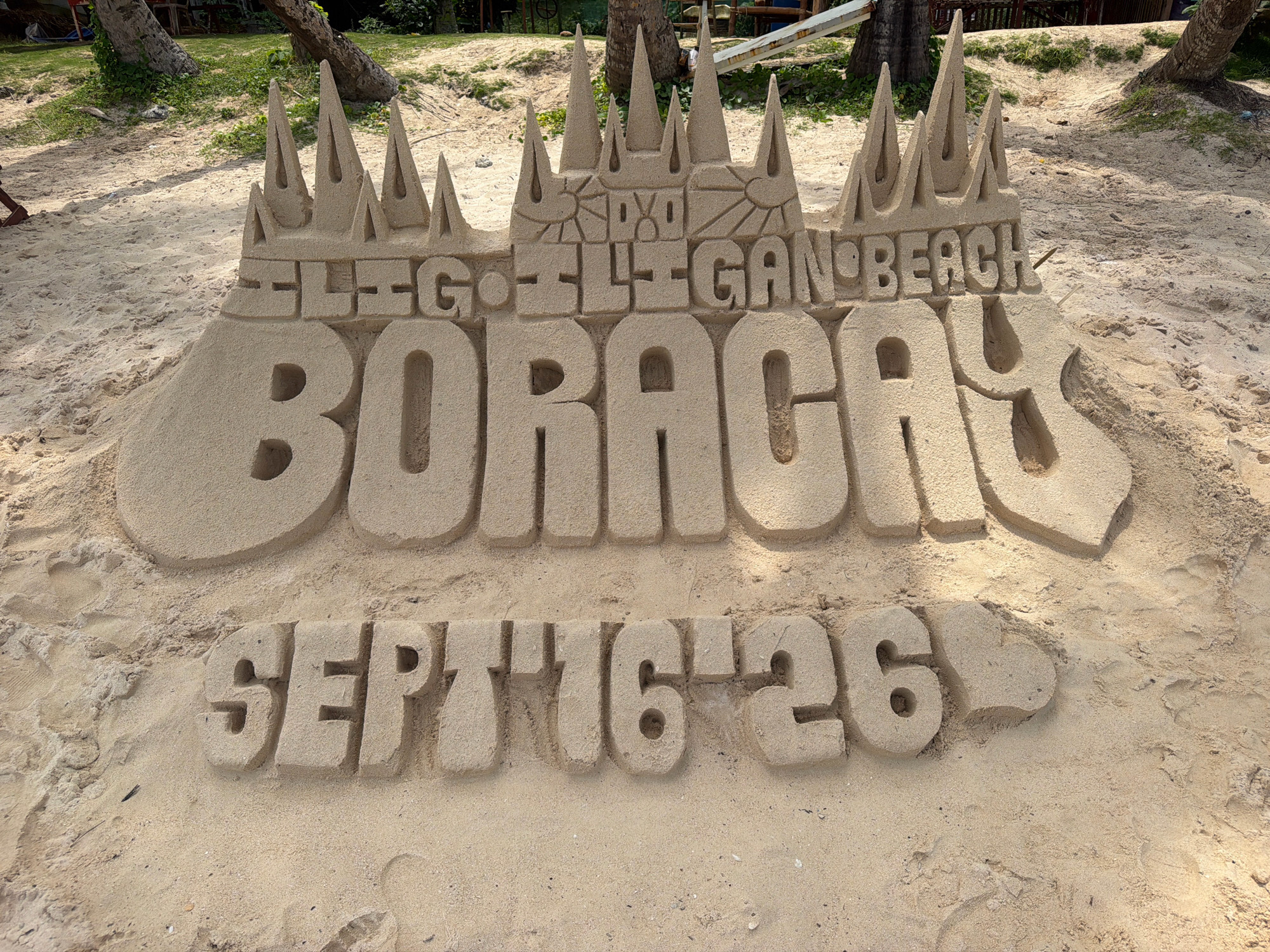 The image shows a sand sculpture on a sandy beach, likely in Boracay, Malay, Philippines, given the text on the sculpture. The sculpture is shaped like a castle with spires at the top. In the center of the castle, the words "ELEGANT" and "BEACH" are carved into the sand. Below this, the word "BORACAY" is carved in large, prominent letters. At the bottom, the dates "SEPT 16-26" are carved, suggesting an event or celebration. The beach is made of light-colored sand. In the background, there are palm trees and some foliage, indicating a tropical environment. The lighting suggests it is daytime, with sunlight casting shadows on the sand. There are no people clearly visible in the frame, though a person's legs and feet are partially visible in the far left background. The sand sculpture appears to be well-maintained and detailed.