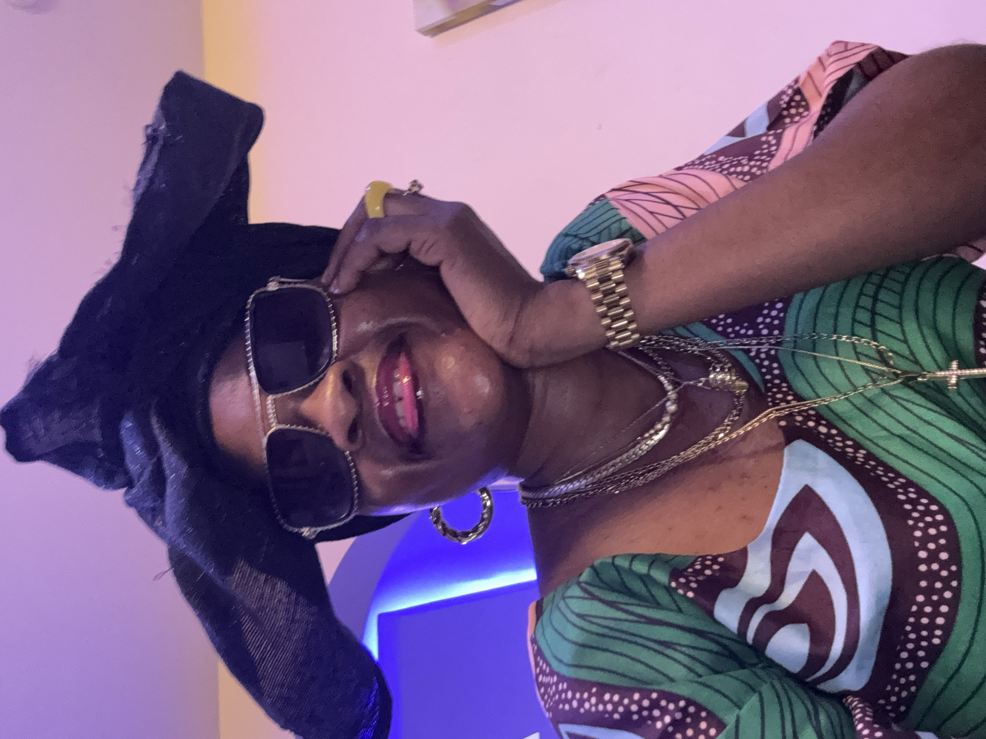 The image features a medium shot of a Black woman wearing a black headwrap, oversized square-shaped sunglasses with silver rims, and a colorful dress with a green, blue, and brown patterned design. She is adorned with several gold necklaces and bracelets, including a chunky chain and a watch. Her left hand is resting on her forehead and cheek, and she has a yellow ring on her right index finger. She has on bright pink lipstick and appears to be smiling or smirking. The background is softly lit with purple and yellow hues, suggesting an indoor setting with artificial lighting. The image is oriented sideways. The woman's attire and jewelry suggest a celebratory or stylish occasion. The provided location context indicates this image was taken in Ajara, Nigeria. No visible text is present in the image.
