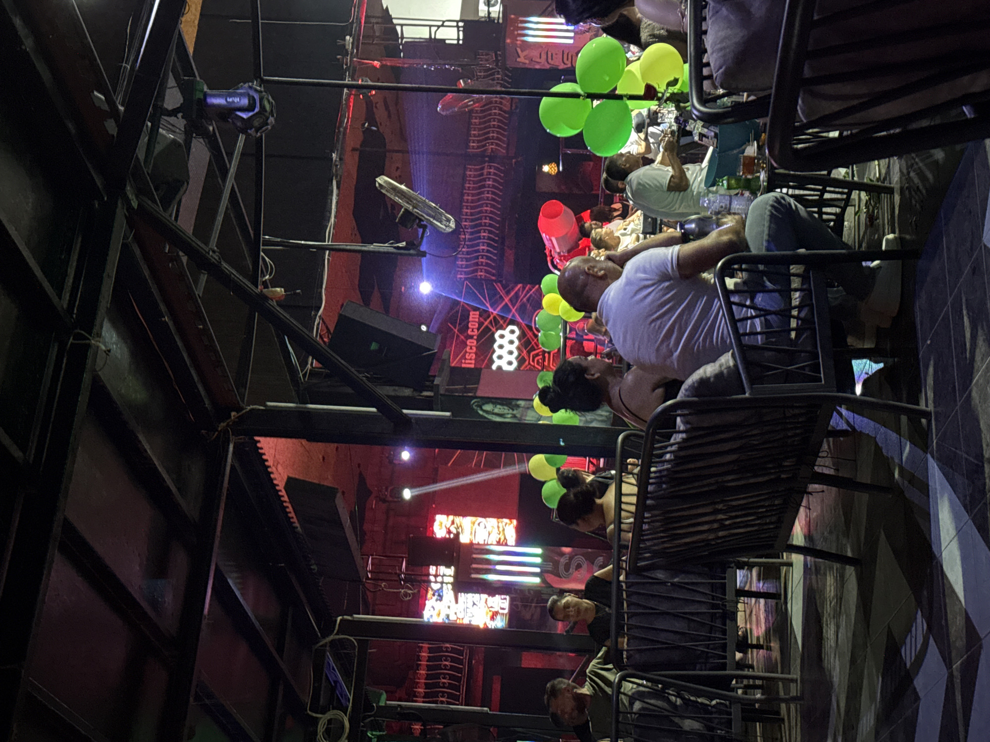 The image depicts a nighttime scene inside a crowded establishment, likely a bar or nightclub, in Alanya, Turkey. A group of people are seated at tables with chairs and sofas, some engaging in conversation or consuming drinks. The atmosphere is dimly lit with colored lights illuminating the space, featuring red and blue hues from stage lighting and spotlights. Balloons in green and yellow are visible, some clustered together. The architecture suggests an open-air or semi-open setting with a metal roof structure and industrial-style beams. In the background, a stage area with lights and what appears to be a screen or backdrop displaying text and graphics, including "lisco.com" and a hexagonal pattern, is visible. The floor is tiled with a geometric pattern. Several individuals are captured in various poses, some looking towards the stage, others interacting with each other. The overall impression is that of an active entertainment venue during operating hours.