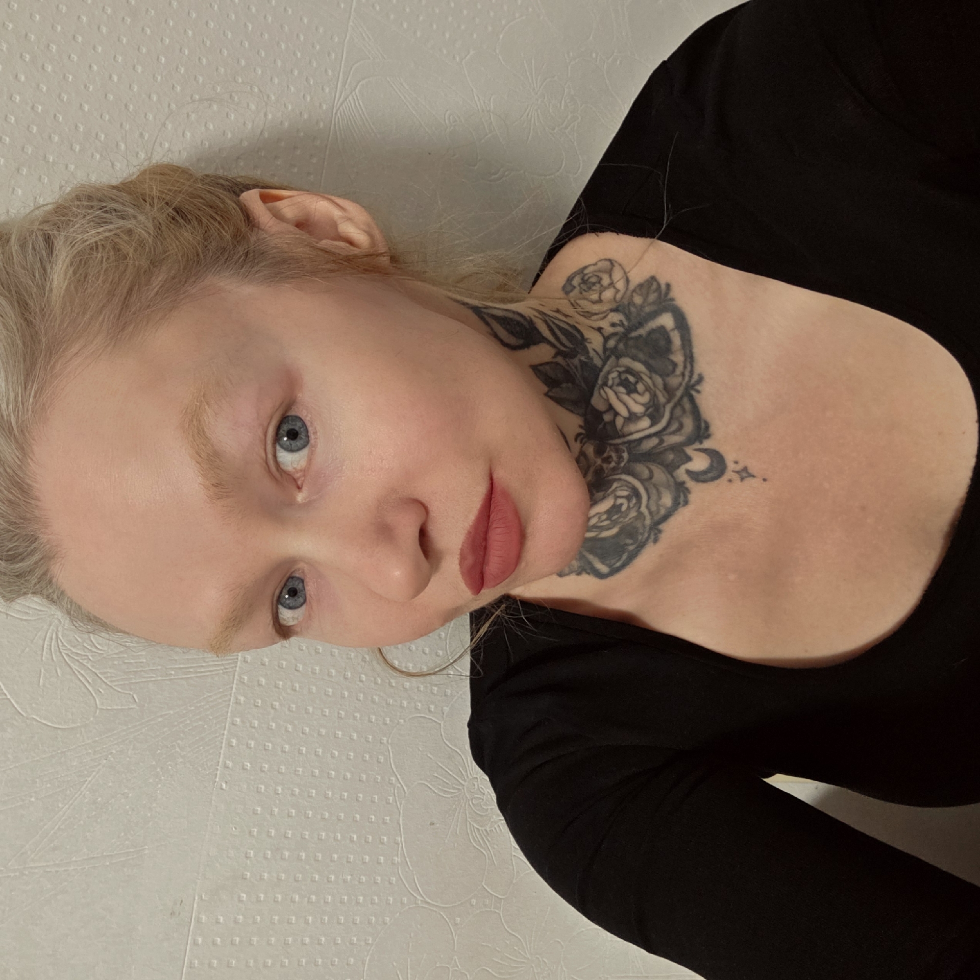 A light-skinned female with blonde hair and blue eyes is lying on a textured white surface, possibly a bed or wall. She is wearing a black top. A detailed floral tattoo is visible on her neck. The location is not identifiable as Saint Petersburg, Russia, based on the information in the image.