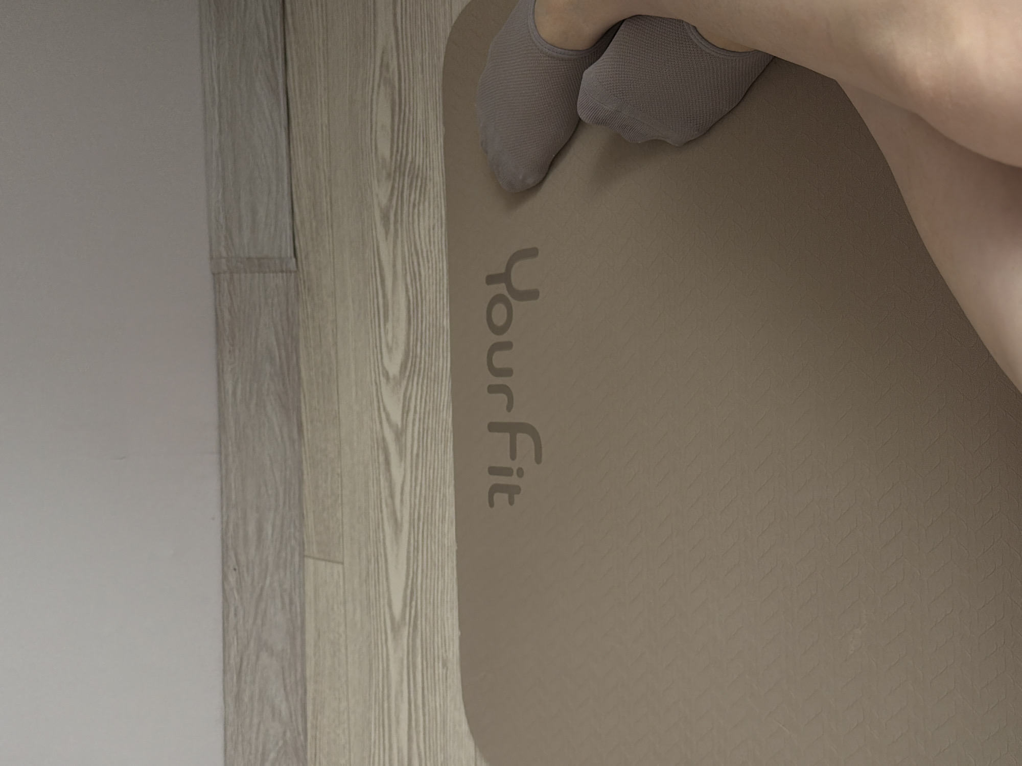 The image displays a close-up, high-angle view of a person's lower legs and feet positioned on a light brown exercise mat. The person is wearing light gray, textured no-show socks, with their feet bent, suggesting they are either sitting or reclining on the mat.
The main subject is a beige or light brown yoga or exercise mat, which features a subtle, repetitive geometric pattern across its surface. Prominently displayed on the mat, oriented vertically from bottom to top, is the brand name "YourFit" in a slightly darker brown or gray font. The letters are rounded and sans-serif.
To the left of the mat, a section of light-colored wooden flooring with visible grain patterns is apparent, indicating an indoor setting. Adjacent to the wooden floor, on the far left, a plain, light-colored wall is visible, possibly white or light gray.
The scene suggests an activity related to exercise, yoga, or stretching, given the presence of the mat. The lighting appears soft and even, without harsh shadows, contributing to a calm and neutral atmosphere. The overall color palette is subdued, consisting mainly of various shades of gray, brown, and beige. The specific time of day is not discernible, nor are any strong emotional cues. The setting is clearly indoors, likely a home or a personal exercise space in Reshetnikovo, Russia.