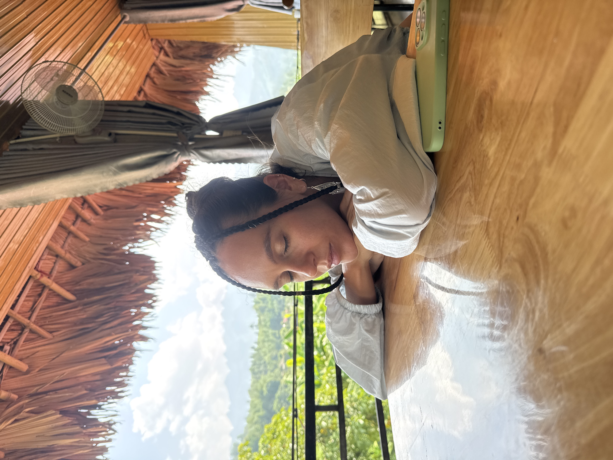 A woman with dark hair braided near her face, wearing a light-colored top, is resting her head on a wooden table. A green phone case is near her head on the table. In the background, outside a window or opening, there are green trees and a cloudy sky. Overhead, there is a thatched roof and a ceiling fan. The scene appears to be indoors in Mậu Duệ Commune, Vietnam.