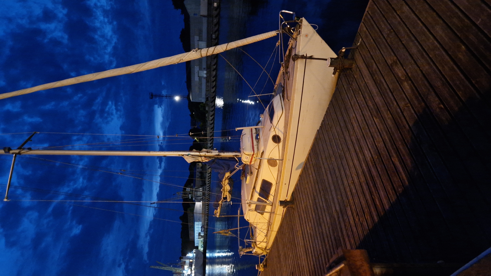 A white sailboat is moored at a wooden pier at dusk or night. The sky is a deep blue with dramatic clouds. The water is dark and reflects lights from the distant shore. In the background, there are industrial buildings, cranes, and lights, suggesting a port or harbor. The sailboat has a mast, rigging, and a sail furled. It is docked alongside the pier. The pier itself is made of weathered wooden planks. The scene evokes a sense of calm and quiet at the end of the day. The specific location in Liepaja, Latvia, cannot be determined from the image alone.
