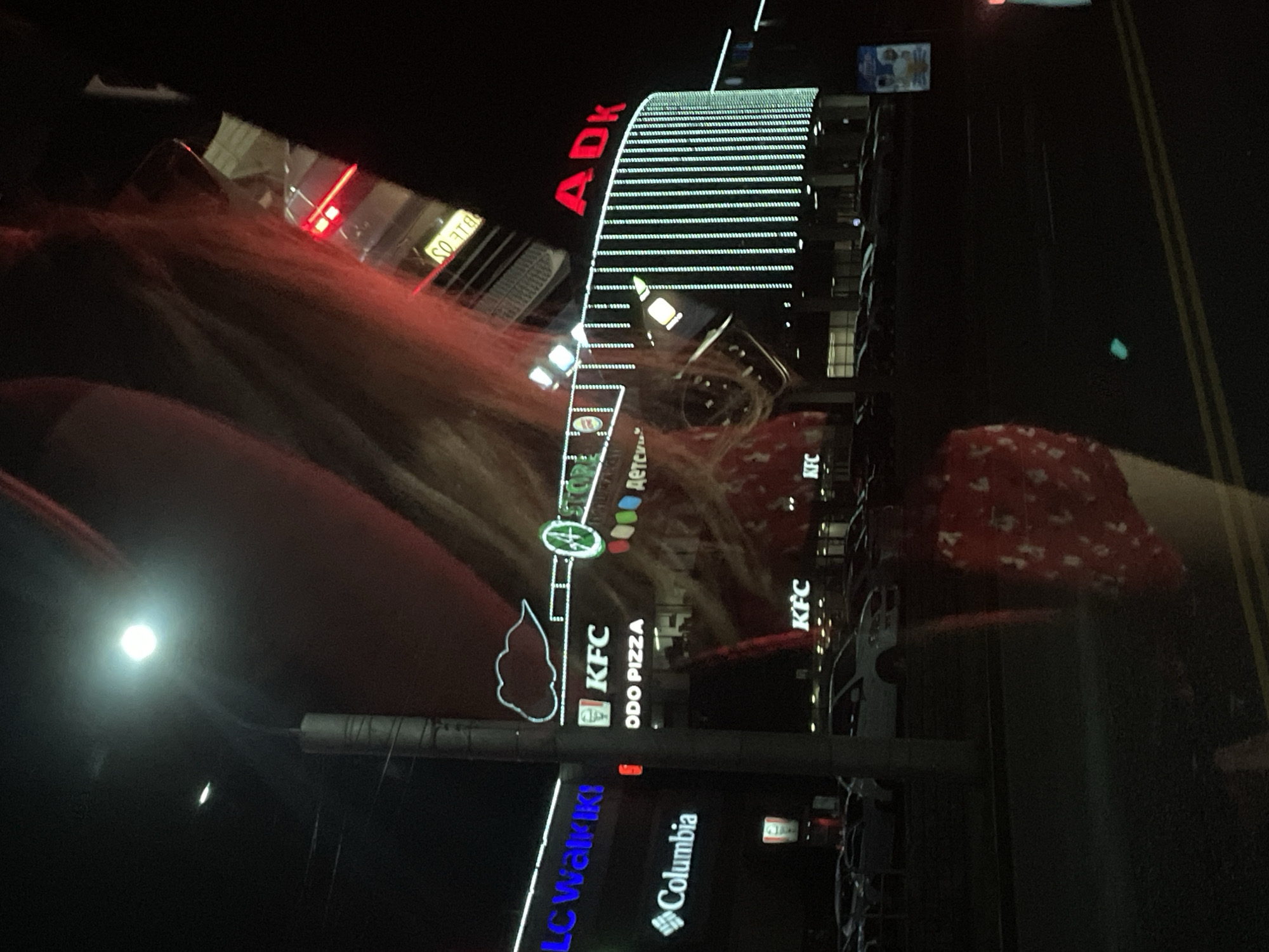 The image shows a night scene with a brightly lit building facade featuring various signs. The building appears to be a commercial establishment, possibly a shopping center or a complex with multiple businesses.
Prominently displayed on the building is a large, curved sign with red lettering that reads "ADK". Below this, a section of the building is covered in horizontal lines of white lights. Further down, there are several illuminated signs for businesses. These include "KFC", "ODO PIZZA", and "LC WALKIKI". Another sign reads "Columbia" with its distinctive logo. One sign, with a green circle containing the number "4", is also visible.
The scene is set at night, indicated by the darkness of the sky and the artificial illumination from the building and streetlights. The lighting is strong, creating reflections on what appears to be a glass surface in the foreground, possibly a car windshield. A blurry, reddish-brown mass is visible in the foreground, which seems to be hair. There are also some people present, though they are largely out of focus and indistinct. The overall impression is one of a vibrant urban environment at night, with commercial activity evident.
The visible text "ADK", "KFC", "ODO PIZZA", "LC WALKIKI", and "Columbia" suggests a retail or entertainment complex. The location context provided is Almaty, Kazakhstan. The weather is not discernible from the image.