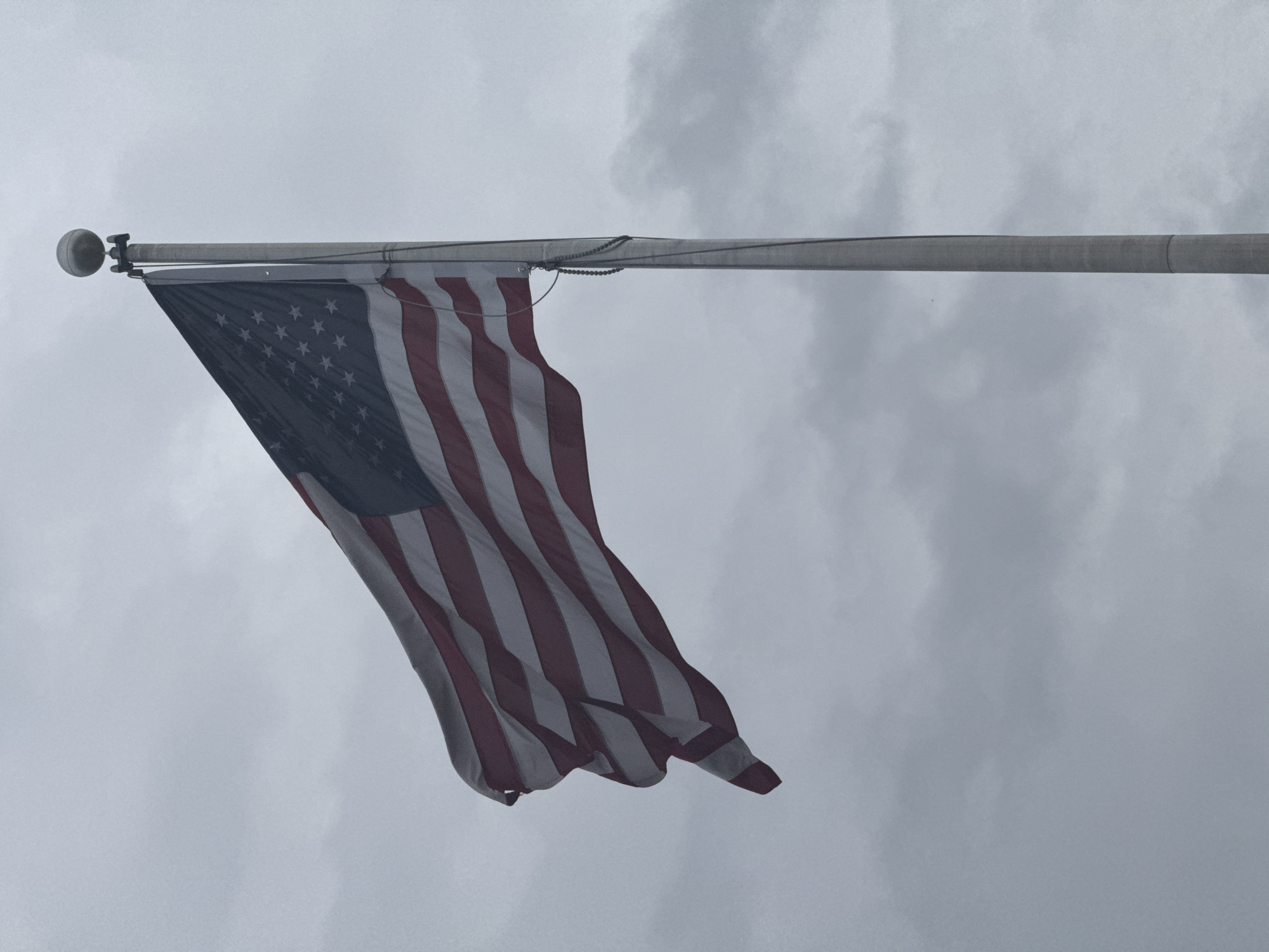 The image shows an American flag hanging from a flagpole against an overcast sky. The flag is waving in the wind, displaying the stars and stripes. The flagpole is a gray metal pole with a spherical top. The environment appears to be outdoors, possibly in Mountain Park, United States of America, based on the prompt. No people or interactions are visible in the image.
