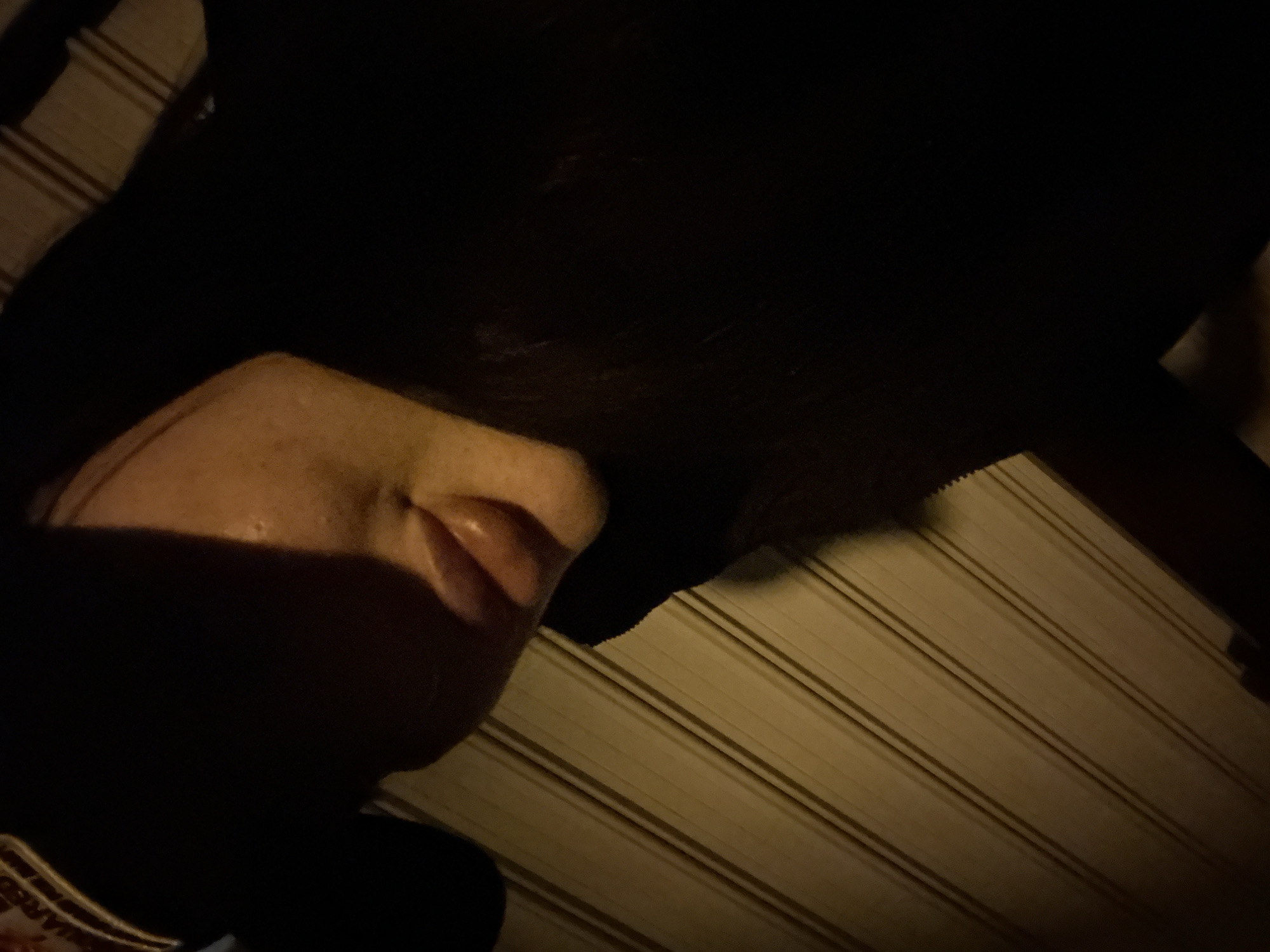 A low-angle, obscured view depicts part of a person's face – primarily the cheek and lips – in shadow, against a background of horizontally oriented lines, possibly corrugated metal, under dim lighting conditions. The location cannot be ascertained from the image.