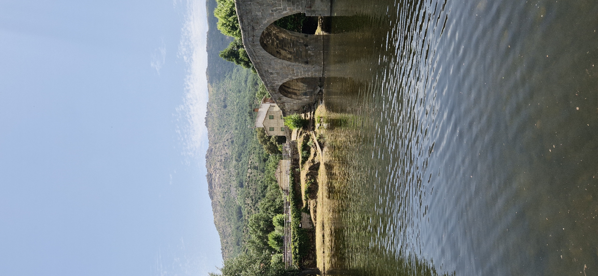 The image shows a scenic view of a river flowing beside a hilly, tree-covered landscape. A stone bridge with a prominent archway spans across the scene, casting a reflection on the water's surface. Below the bridge, a small, light-colored building with green-trimmed windows is nestled amidst the vegetation. The terrain is characterized by steep, verdant hills and terraced slopes, suggesting a rural or natural setting. The water appears calm with ripples, reflecting the sky and the bridge. The overall mood is serene and picturesque. The time of day appears to be daytime, with bright sunlight illuminating the scene. The weather seems clear, indicated by the blue sky. There is no visible text in the image.