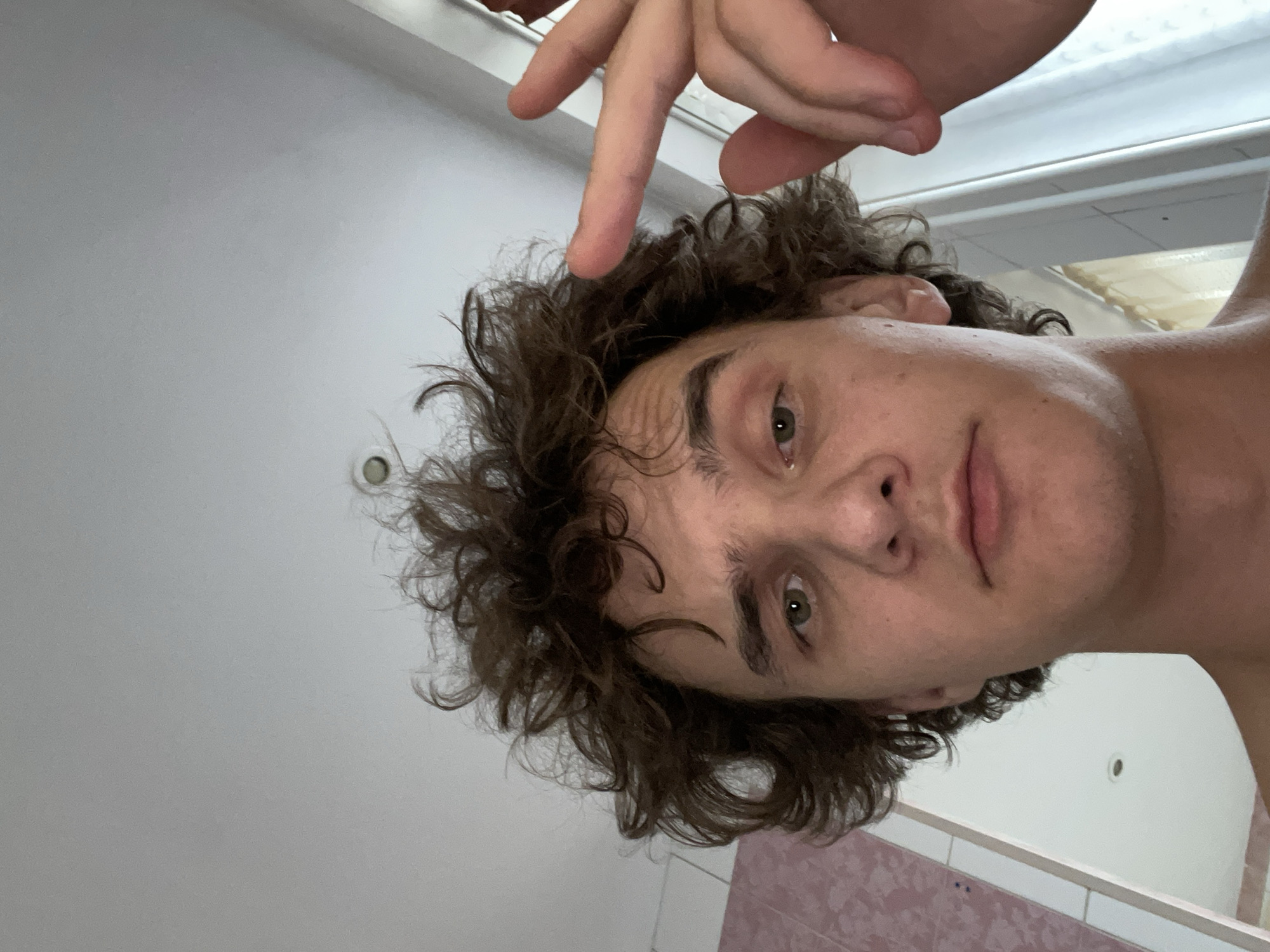 This is a selfie taken by a young person with curly brown hair and green eyes. They are pointing their right index finger towards their forehead. The person appears to be in a bathroom, with white tiled walls and a pink patterned surface visible at the bottom of the frame. A round light fixture is visible on the wall in the background. The lighting suggests it might be daytime, but no external weather cues are present. The expression on the person's face is neutral. No text is visible in the image. The setting of Krasnoyarsk, Russia is provided as context.