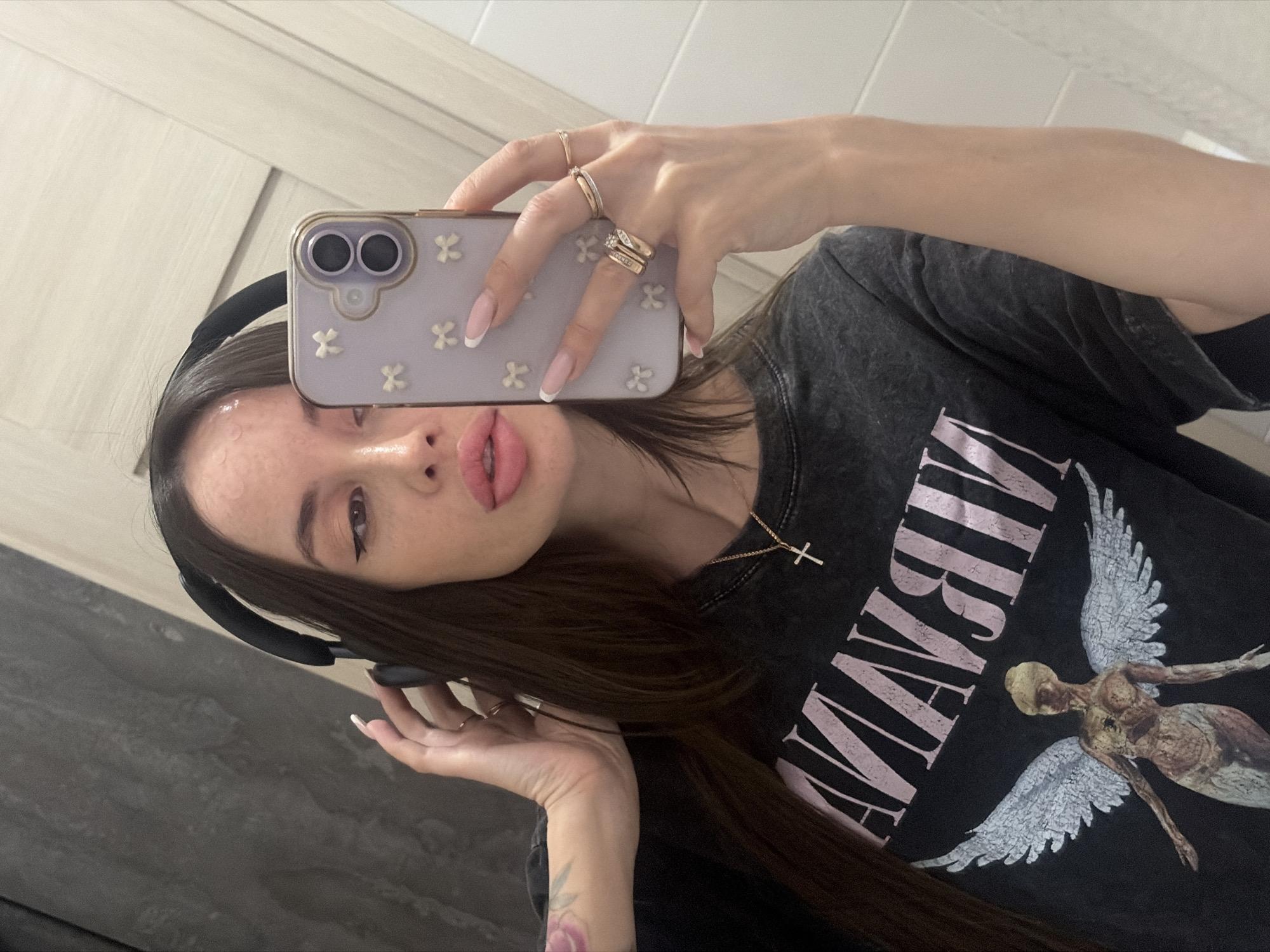 A young woman is taking a selfie in front of a bathroom mirror. She is holding up a purple phone with a floral pattern, obscuring part of her face. She is wearing a black Nirvana t-shirt and a gold necklace with a cross pendant. She also has headphones on. Her hair is long and dark brown. She is wearing multiple rings on her fingers and her nails are manicured with a French tip. The background consists of a light-colored door with recessed panels and a dark gray wall. There is no information in the image to support the conclusion that this photo was taken in the city of Saint Petersburg, Russia.