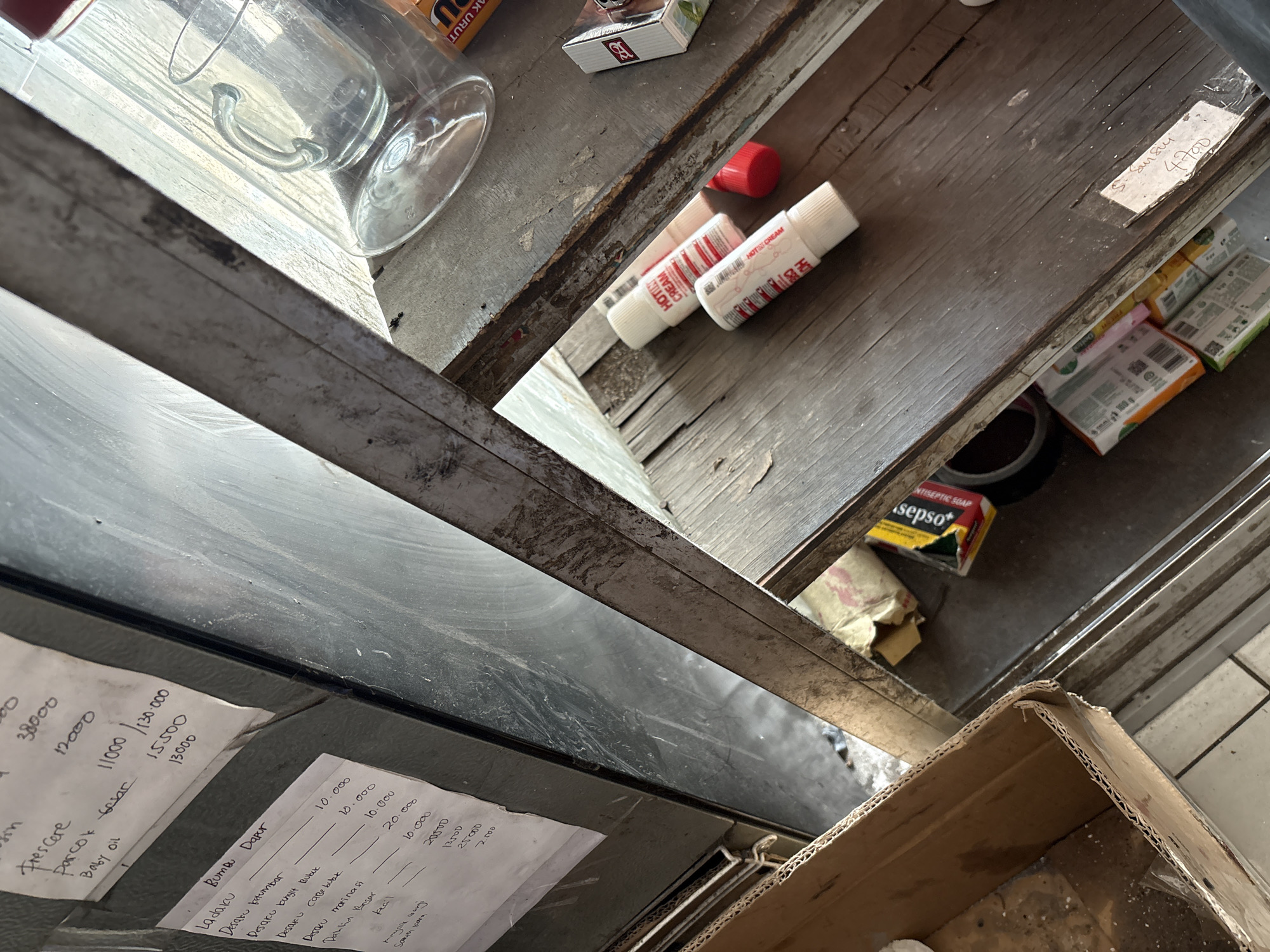 The image depicts an indoor scene, likely a storage area or workshop, with a focus on a wooden shelf laden with various items. The foreground shows a cardboard box. On the shelf, two tubes of "HOT STICK CREAM" are visible, alongside a red cap. A glass mug and saucer are also present on the shelf. To the left, a piece of paper with handwritten text and numbers, appearing to be a list or receipt, is attached to a metal surface. Further to the right, a small box of what looks like antiseptic soap is present, along with other unidentifiable items. The overall environment appears somewhat cluttered and aged, with visible wear and tear on the wooden surfaces and metal fixtures. There are no people visible in the image, and no discernible actions or interactions are taking place. The lighting suggests it is daytime, with sunlight casting shadows.