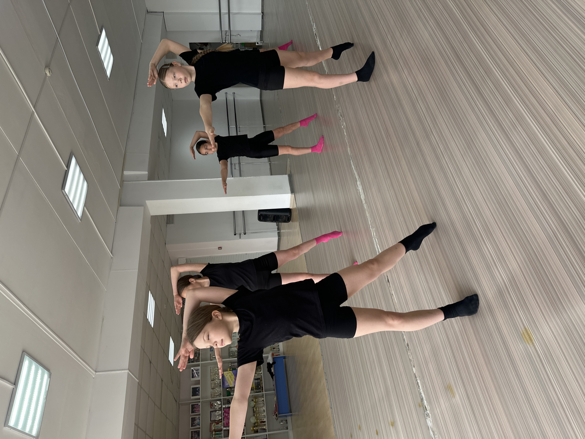 Three young girls are practicing a dance routine in a dance studio. The studio has a reflective wall, creating the impression of six figures. The girls are wearing black shirts and shorts, with socks of varying colors (black and pink). They are extending one arm upward and the other to the side while balancing on one leg. The studio has a light-colored wooden floor and white walls. There are rectangular ceiling lights visible. In the background, near the reflective wall, is a shelf with trophies and awards. The location is likely a dance studio in Votkinsk, Russia, but without explicit geotags or contextual information, this is difficult to confirm with certainty.