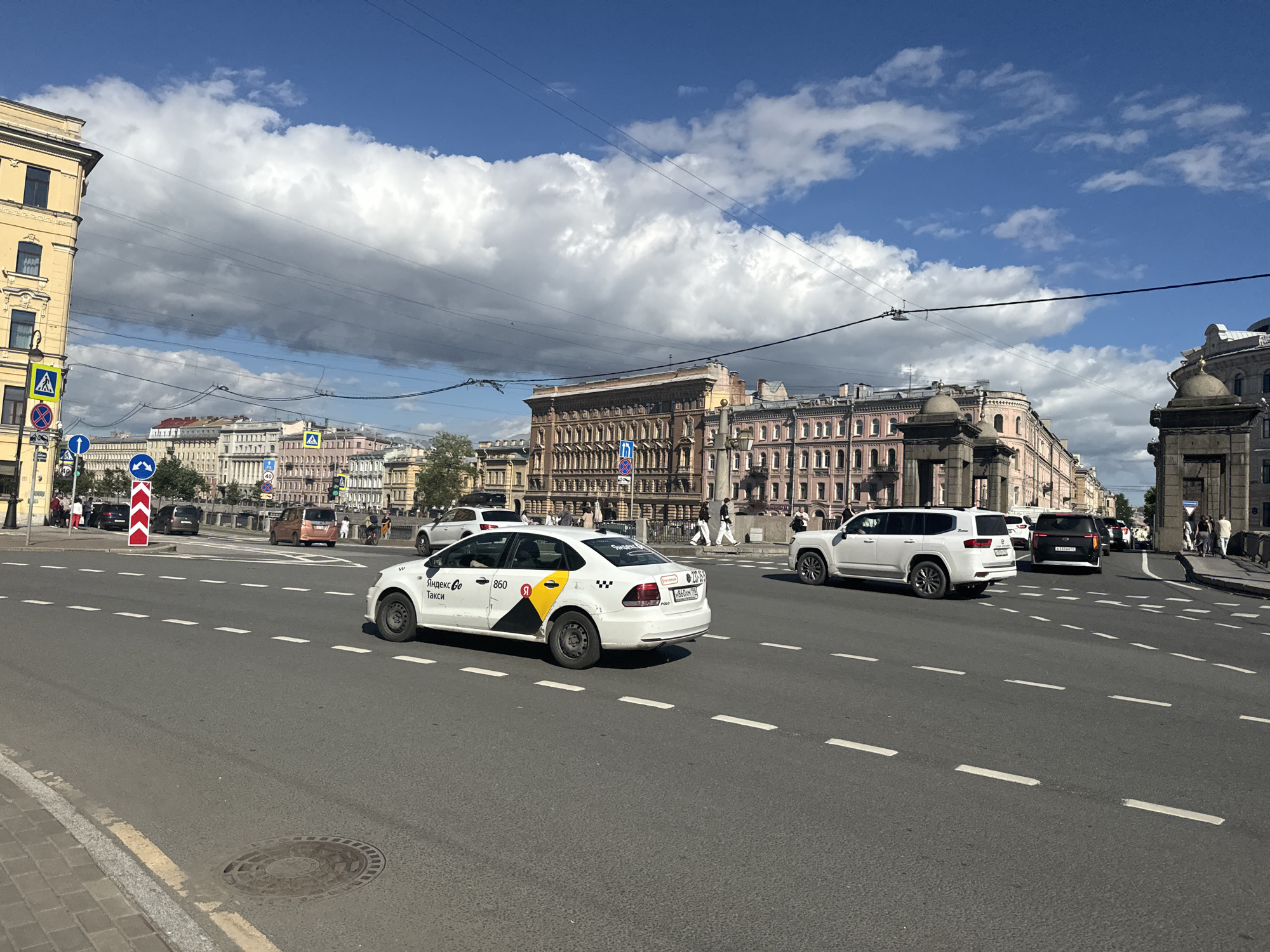 The image shows a street scene in Saint Petersburg, Russia on a sunny day with scattered clouds. A white taxi with "Яндекс Go Такси" text is in the foreground, driving on a multi-lane asphalt road. The taxi has a yellow and black checkered pattern on its rear passenger door. Behind the taxi, other vehicles including a white SUV and a dark SUV are visible. In the background, ornate buildings with classical architecture line the street, characteristic of Saint Petersburg. Pedestrians are walking on the sidewalk in front of the buildings. Traffic lights and road signs are present, indicating an urban environment. The time of day appears to be midday due to the bright sunlight and shadows. The overall scene depicts a typical day in a bustling city.