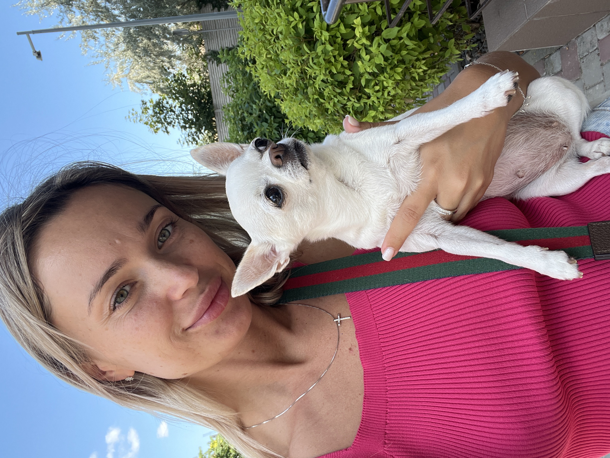 A woman with blonde hair and green eyes, wearing a bright pink top, is taking a selfie outdoors while holding a small white chihuahua. She has a silver necklace with a cross pendant. The chihuahua is looking to the right of the frame. The background consists of green bushes, a fence, and a clear blue sky with some clouds. A street lamp is visible in the distance. The scene appears to be in Kryzhanivka, Ukraine, a coastal village near Odesa.