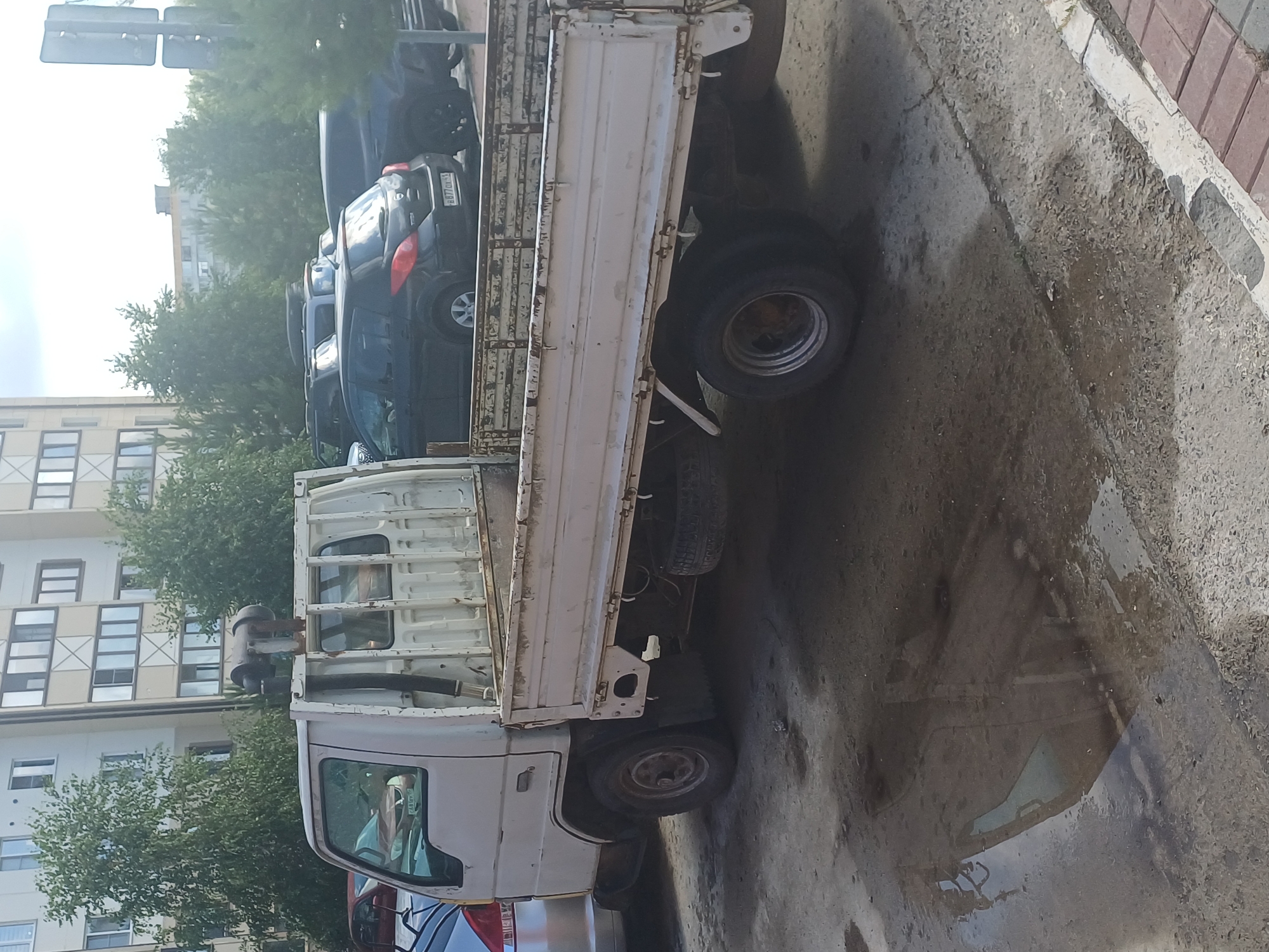 In Elitnyi, Russia, a white flatbed truck is parked on a paved surface near a multi-story apartment building. The truck appears weathered, with visible rust on its bed and other components. A spare tire is secured underneath the truck bed. Other vehicles, including dark-colored cars and SUVs, are parked behind the flatbed truck. Trees line the area, and the sky is overcast. The ground surface shows signs of wetness, possibly from recent rain, creating reflections of the surrounding environment.
