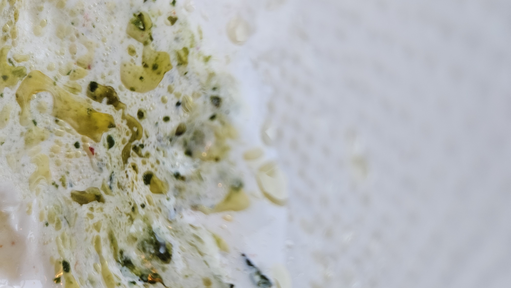 The image is a close-up, abstract shot of a creamy white substance with streaks of green and yellow oil, appearing to be a food item. The substance has a foamy texture and some small, dark specks. The bottom portion of the image is out of focus, showing a blurry, pale surface. There is no discernible location in L'Île-Rousse, France.