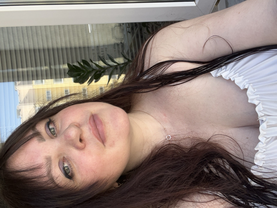 The image displays a close-up, chest-up portrait of a female subject, reportedly located in the city of Noviy, Russia. The subject has fair skin, dark brown hair with bangs, and prominent strands that appear damp, falling over her shoulders and chest. Her eyes are green/hazel, enhanced with dark eyeliner and purple eyeshadow. Her lips are lightly tinted with a pink or nude color and are slightly parted. Small gold-colored hoop earrings are visible in her left ear, and a delicate gold-colored necklace with a circular pendant and cascading small beads is worn around her neck, resting on her chest. A reddish mark is present on her neck below her left ear. She is attired in a light-colored, possibly white, garment with a ruffled or elasticized neckline. The background reveals an urban environment through a large window with partially closed vertical blinds. Outside the window, multiple residential buildings, one appearing light yellow or beige, are visible under a clear blue sky. A green potted plant with dark, ovate leaves is positioned near the window. The scene is illuminated by bright, natural daylight.