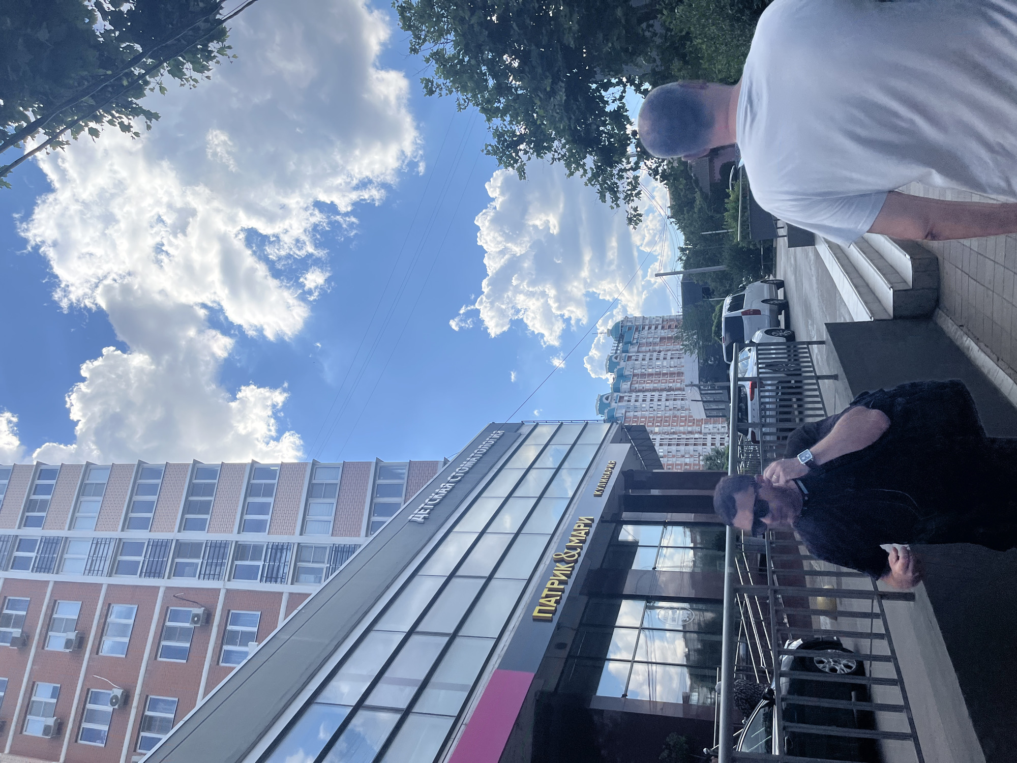 In Krasnodar, Russia, on a clear day with fluffy white clouds dotting the bright blue sky, two men are visible in the foreground. One man, wearing a white t-shirt, is seen from behind, his head and shoulders angled away from the camera. The other man, dressed in black, is also partially visible from the side, wearing sunglasses and holding a white object, possibly a phone or a receipt. They are standing on a paved area next to a modern building with a glass facade.
The building has prominent signage. On the glass facade, text in golden letters reads "ПАТРИК & МАРИ" (PATRICK & MARI), and above it, on a darker panel, the text "ДЕТСКАЯ СТОМАТОЛОГИЯ" (CHILDREN'S DENTISTRY) is visible in white letters. The building's architecture features a mix of glass, dark metal framing, and sections of light-colored brick.
In the background, several other buildings are visible, including a tall residential building with a blue and white facade. Cars are parked along the street, and trees line the background, suggesting an urban or suburban environment. The sunlight indicates it is daytime, likely late morning or afternoon. The overall atmosphere appears casual and everyday.