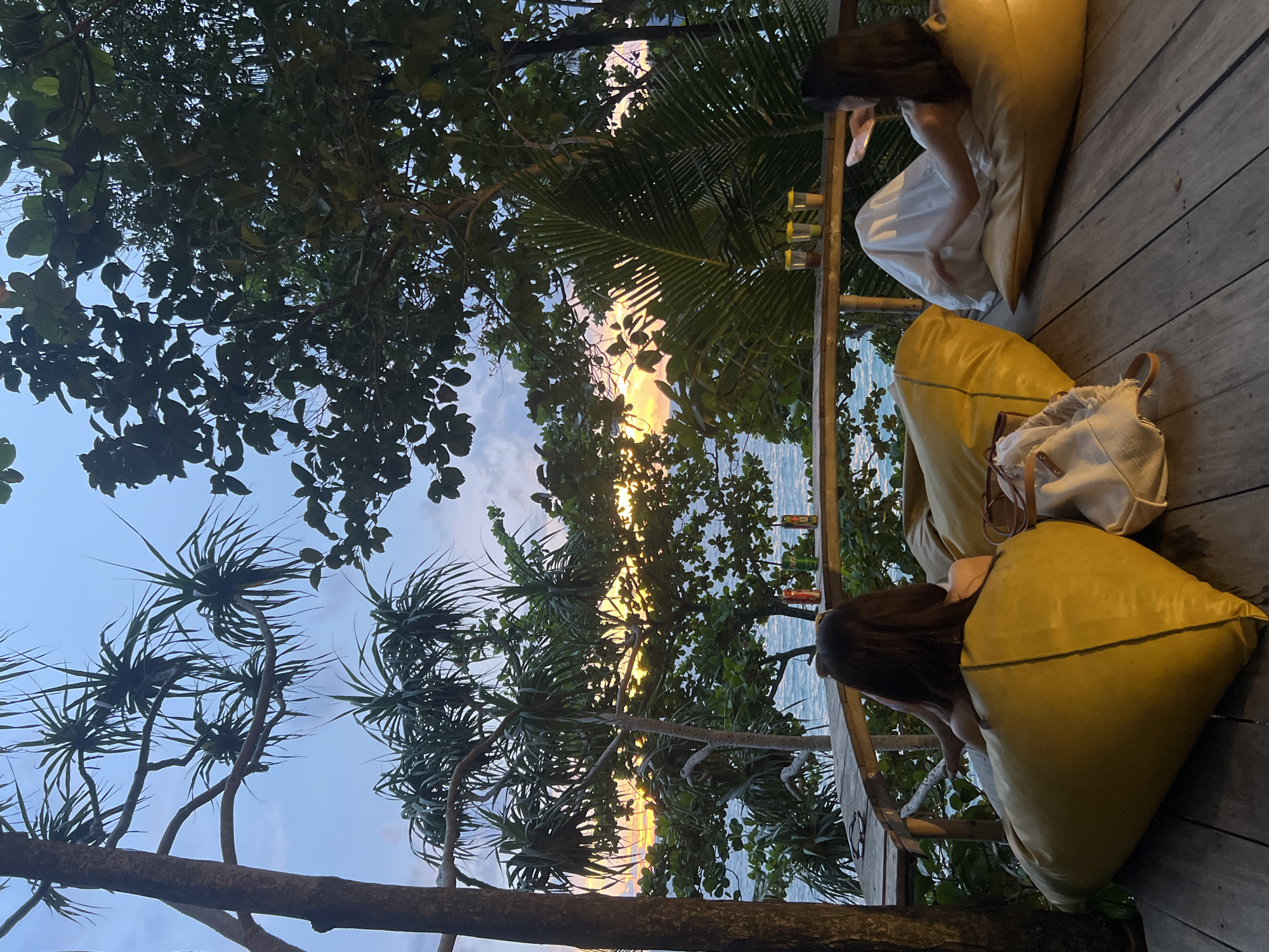 Two individuals are seated on large yellow beanbag chairs on a wooden deck overlooking the ocean at sunset in Karon, Thailand. The person on the left, facing away from the camera, has dark hair and is wearing a light-colored garment. The person on the right, also facing away from the camera, is wearing a white dress and has long dark hair. Between them on the deck are two bags, one a light-colored tote bag. In the background, the ocean is visible, with a sunset casting a warm glow on the horizon. Lush green trees and foliage surround the deck, with palm-like plants in the foreground and other leafy trees creating a natural canopy. A wooden railing separates the deck from the ocean view. On the railing in the distance, there are several glasses and cans of beverages. The sky is a mix of blue and sunset colors.