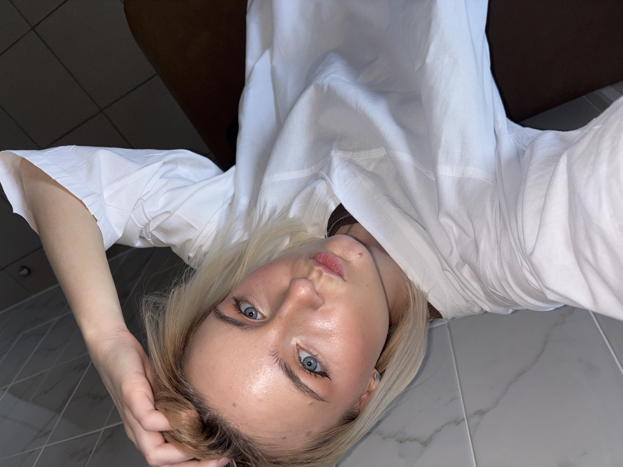 The image displays an indoor, close-up selfie of a young woman with blonde hair and blue eyes, captured in what is understood to be Saint Petersburg, Russia. She is positioned with her head tilted, looking directly into the camera with a composed, neutral expression and slightly parted, natural pink lips. She wears a white collared shirt or dress, and a delicate silver chain necklace is visible around her neck, along with a small stud earring in her right ear. Her right hand gently touches her hair near her head.
Her face is well-lit, highlighting smooth skin with a subtle, dewy sheen on her forehead, nose, and chin. She has light eye makeup, featuring winged eyeliner that accentuates her prominent blue eyes. The background suggests a domestic setting, with a grey tiled floor, characterized by white grout lines and a subtle marble pattern on some tiles, occupying the lower portion of the frame. A dark brown, undefined object or surface is present in the upper background, possibly furniture or part of a wall. The lighting appears to be ambient indoor light, casting an even glow on her face. There is no visible text in the image.
