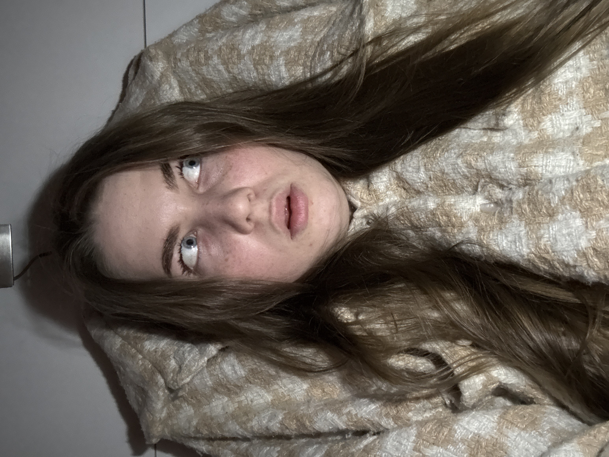The media file presents a candid, close-up portrait of a young woman lying down. She has long, straight, medium-brown hair that spreads out around her head and over a patterned garment. Her complexion is fair, with visible freckles or redness, and she possesses striking light blue or grey eyes. Her gaze is directed upwards and to the side, with her mouth slightly open, creating an expression that could be interpreted as contemplative, tired, or simply captured in a moment of rest.
She is dressed in or lying on a thick, textured garment, possibly a jacket or blanket, featuring a woven pattern in shades of cream, beige, and light brown, resembling a houndstooth or check design. The background is a plain, light grey or white wall or surface, suggesting an indoor setting. A small, metallic, or reflective object is partially visible along the far-left edge of the frame. The lighting is somewhat dim, casting subtle shadows and highlighting the textures of her hair and the fabric. The image is rotated counter-clockwise, creating an unusual, diagonal composition. There is no visible text in the image. The provided location context is Chaikovsky, Russia.