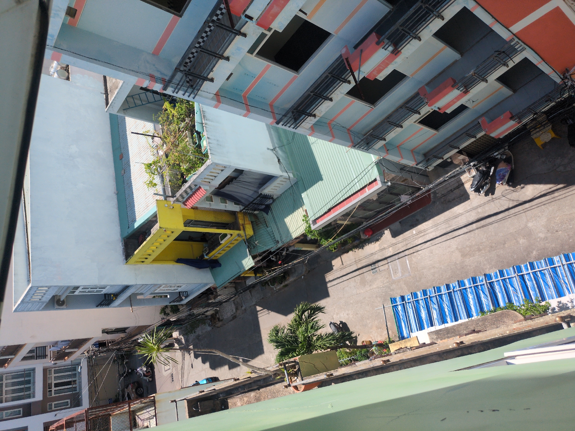 An elevated, high-angle view depicts a narrow, paved street canyon within South Quy Nhon Ward, Vietnam, flanked by multi-story residential and commercial buildings under clear daylight.
On the left side, a light green building, likely the vantage point, frames the foreground. Below it, a series of white and brown buildings with multiple windows and balconies extend along the street. One building exhibits a metallic mesh enclosure. Further down the street, a blue sedan is parked, and a palm tree stands near the curb. A long, blue and white striped tarp or awning is laid flat on the ground along the edge of the street.
The street itself is dark asphalt or concrete, with prominent shadows cast by the buildings and trees, indicating a sunny environment. Overhead, a dense network of black electrical wires crisscrosses the space between buildings. A person on a white scooter is observed driving down the street towards the upper left.
On the right side of the street, a tall, light blue building with horizontal orange/peach stripes and dark metal balcony railings is prominent. Below it, a yellow building with a green corrugated roof is visible, along with multiple parked scooters at street level. Further back, a white building features a rooftop garden with various potted plants and a small blue access ladder. Beyond this, a light-colored building with visible water tanks on its roof is present. The architectural style includes common Vietnamese urban characteristics, such as narrow facades and balconies.