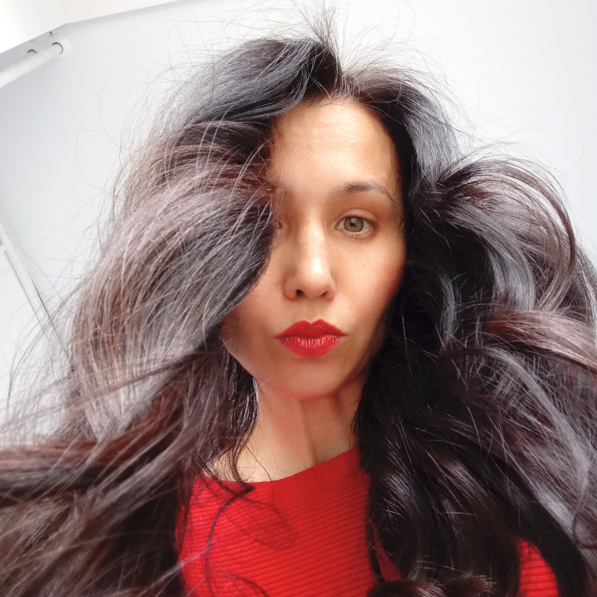 A low-angle close-up captures a woman with flowing, dark hair with graying roots. She is wearing a red top and bright red lipstick. The background is a plain, white ceiling. There is no information available to place the location as Izhevsk, Russia.