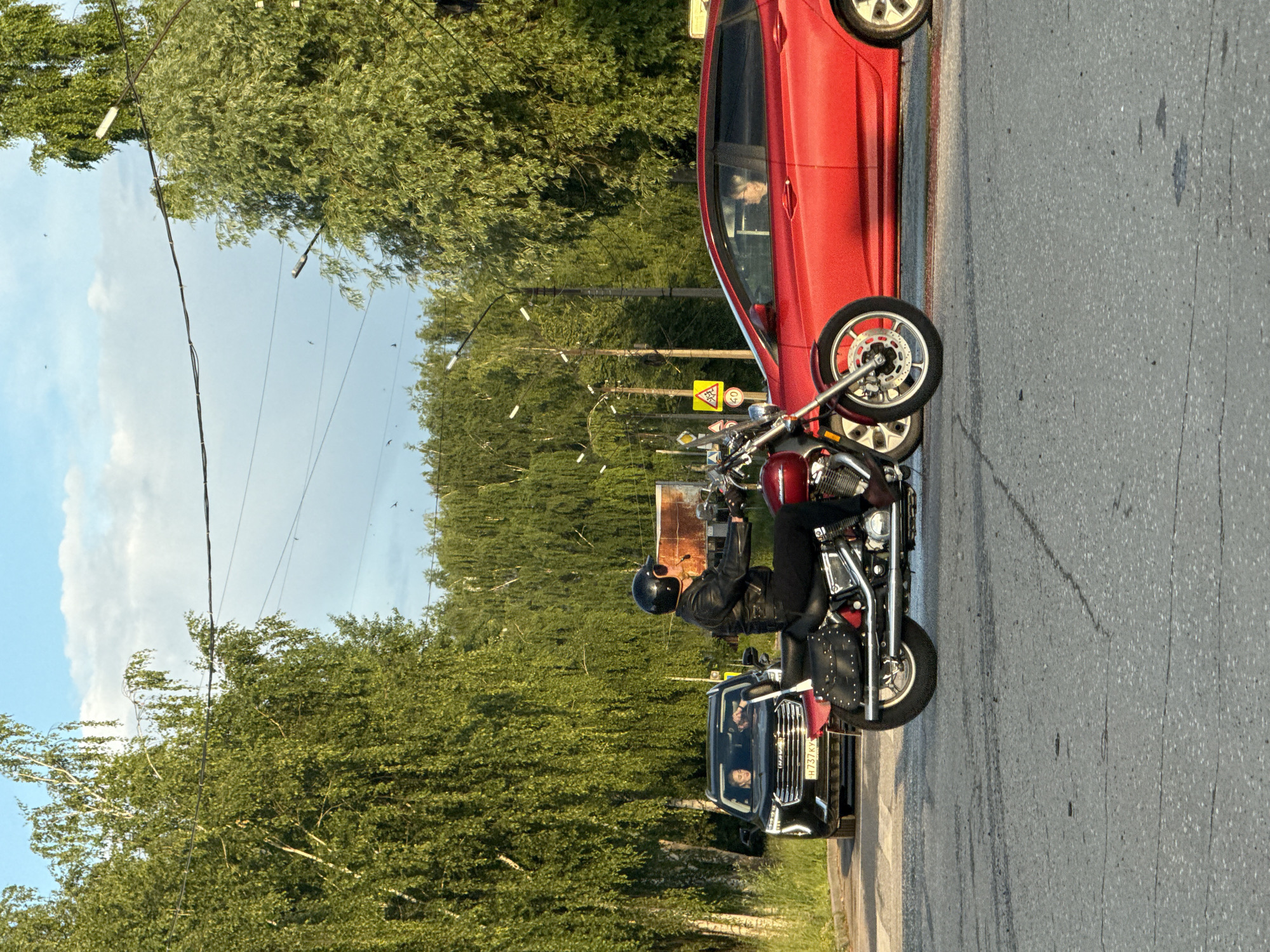 In Votkinsk, Russia, on a paved road surrounded by lush green trees, a person is riding a red motorcycle. The rider is wearing a black leather jacket, black pants, and a black helmet with a visor. The motorcycle has saddlebags on the sides. In the background, a black SUV is stopped, and a red sedan is passing by. The sky is a light blue with scattered clouds. The scene appears to be a typical daytime street view with vehicles and foliage.