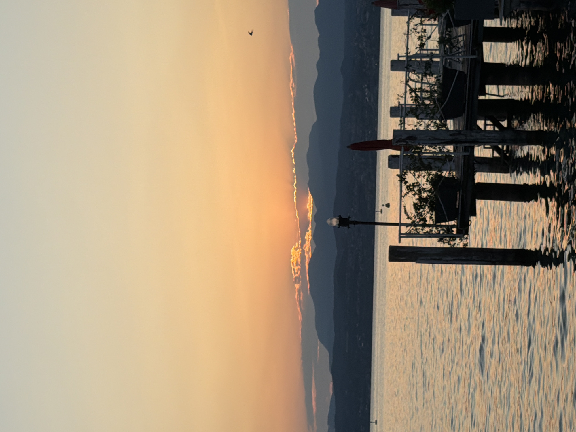 This image depicts a sunset over Lake Garda, viewed from a dock in Sirmione, Italy. The sky is a gradient of warm oranges and soft yellows near the horizon, transitioning to a pale blue at the top. Silhouetted mountains form the distant backdrop, with a thin layer of clouds catching the last rays of sunlight. In the foreground, a wooden dock extends into the water. The dock is supported by pilings and features planters with greenery, a railing, and a lamppost. The water's surface is calm, reflecting the colors of the sky and the structure of the dock. A single bird is visible in flight against the sunset. The overall scene is serene and captures the tranquil beauty of the Italian lakeside at dusk.