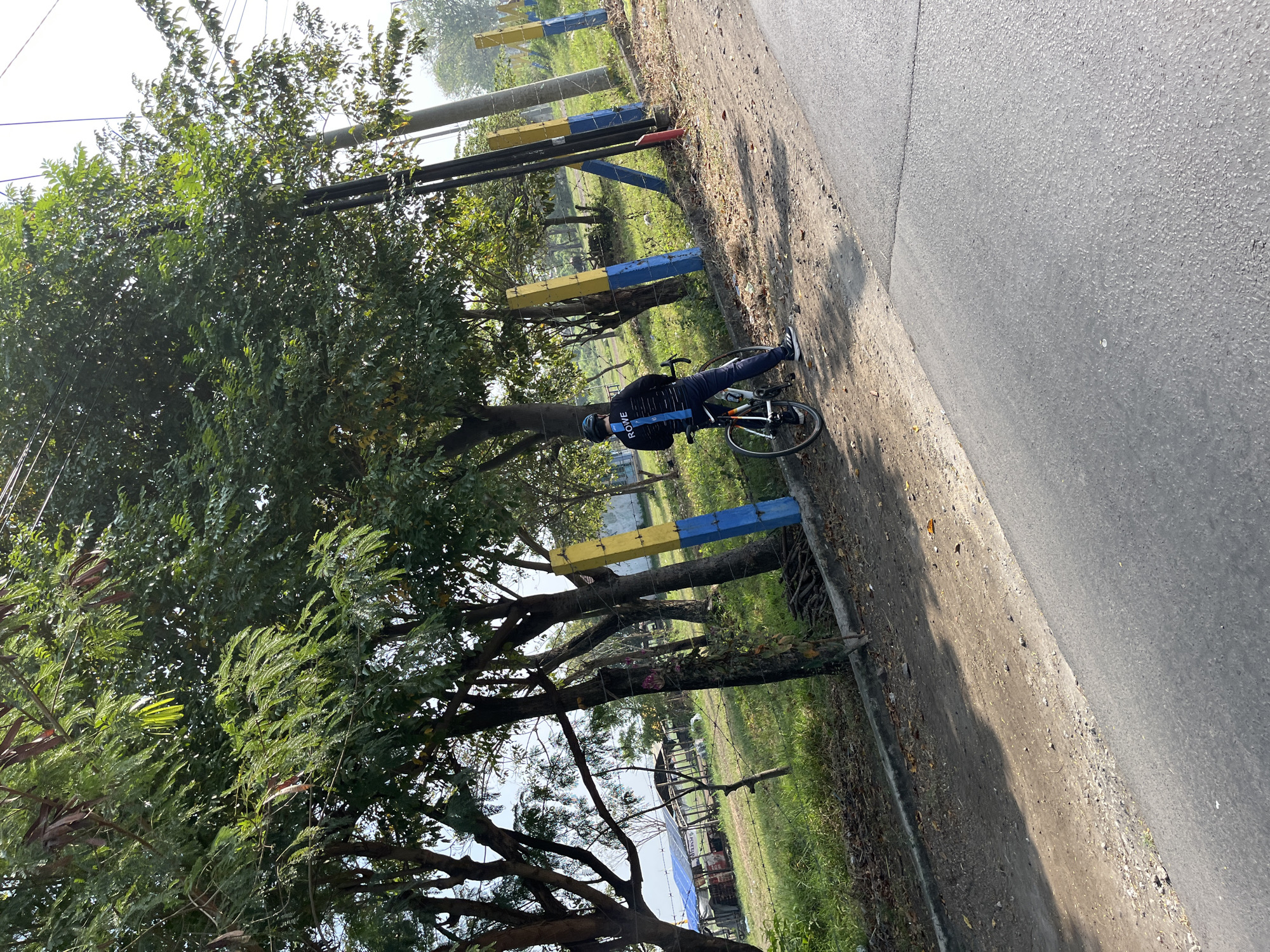 A person is standing next to a bicycle on the side of a paved road in Gempolsari, Indonesia. The person, wearing a black helmet and a black long-sleeved shirt with blue and white stripes on the back, is facing away from the camera and looking towards a grassy area with trees. The bicycle is a road bike with a white frame and orange accents. In the background, there are several poles painted yellow and blue, a fence, and a blue tarpaulin structure. The environment appears to be a rural or semi-rural area with abundant greenery and clear daylight. The road surface is asphalt.