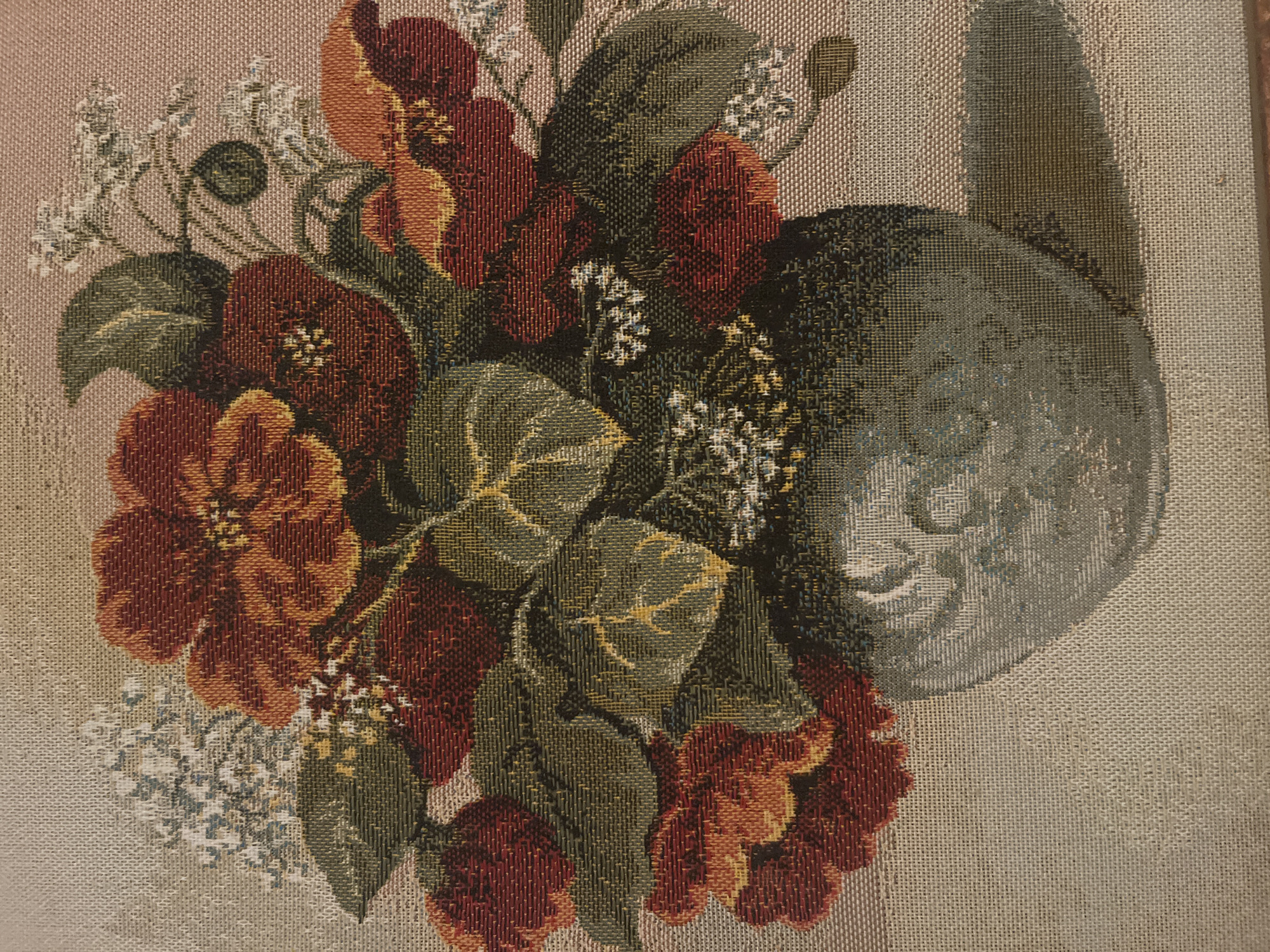 This is a tapestry depicting a floral arrangement in a vase. The flowers are predominantly in shades of red and orange, with some green foliage. The vase appears to be a light gray or blue color with decorative patterns. The background is a muted beige or light pink. The texture of the tapestry is visible, showing the woven threads. There are no people or discernable elements that would suggest a specific location within the city of Parekklisia Community, Cyprus.