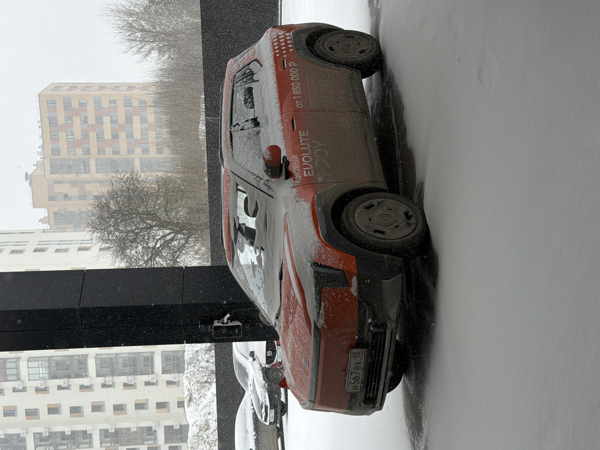The image shows a red Evolute i-JOY car parked on a snowy day. The license plate reads "H567 BX 18." The car has "EVOLUTE i-JOY" written on the side, along with "from 1,850,000 rubles." Buildings are visible in the background, suggesting a cityscape. The location is Izhevsk, Russia.
