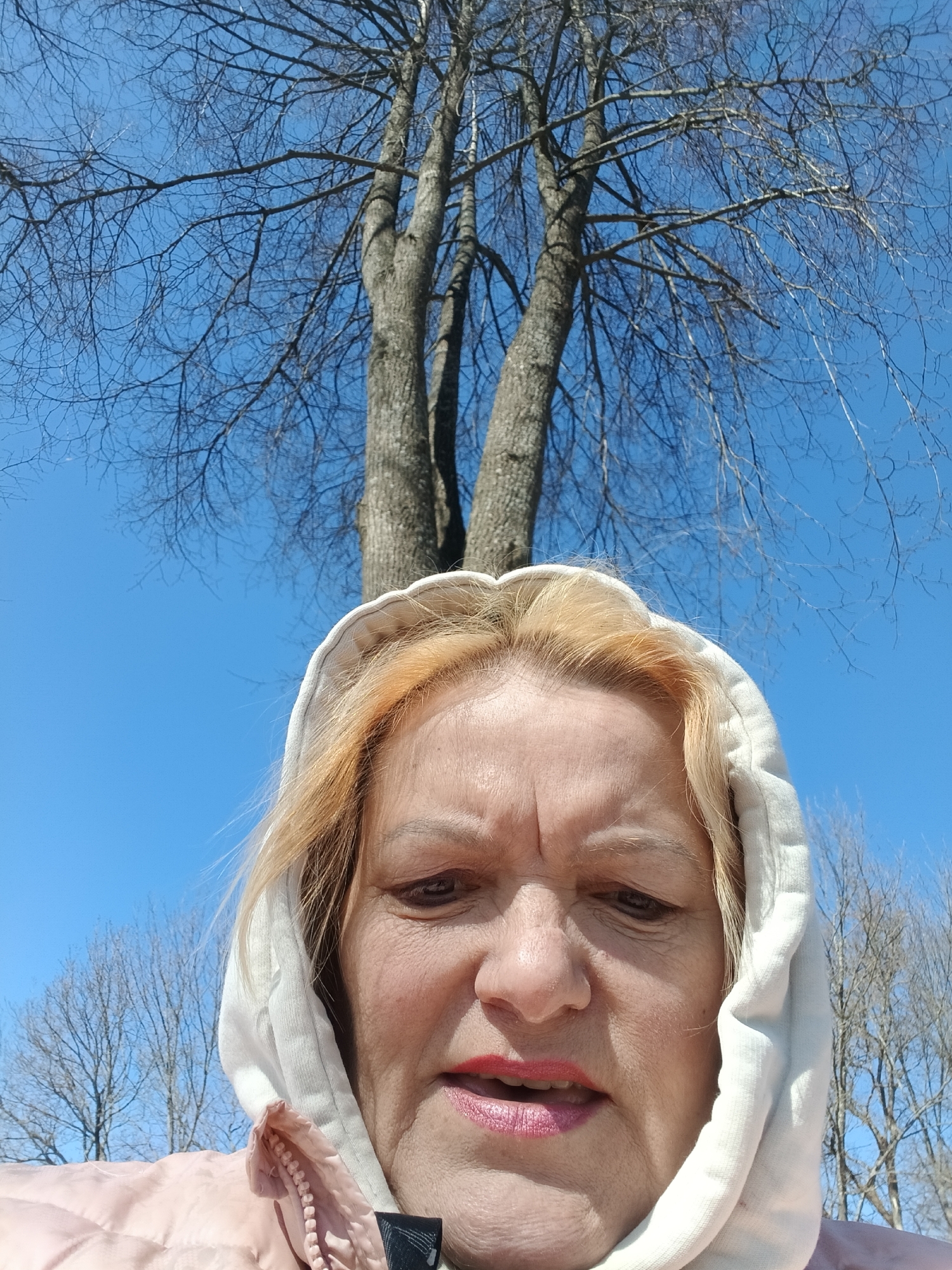 A low-angle outdoor shot captures a middle-aged woman in the foreground against a backdrop of bare trees and a clear blue sky in Красный Маяк, Russia. The woman's head and shoulders fill the lower half of the frame. She has blonde hair, with visible roots, partially covered by a light-colored (possibly white or off-white) hooded sweatshirt. She wears a pink or light salmon-colored puffer jacket underneath the hoodie. Her expression is somewhat pensive or serious, with a slight frown, and her mouth is slightly open, revealing red lipstick.
Above her, the intricate network of bare branches and grey trunks of deciduous trees stands out against a bright, cloudless blue sky. The starkness of the bare branches indicates either late winter or early spring. The lighting is bright, suggesting a sunny day. No visible text is present in the image.