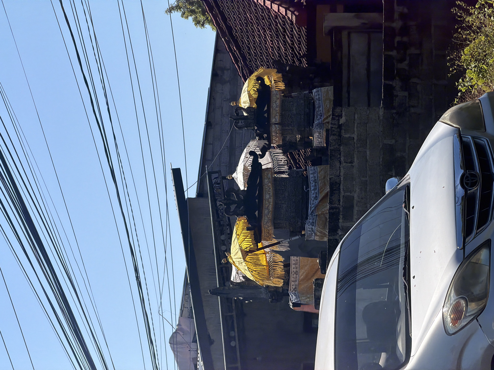 The image shows a street scene in Kubu, Indonesia, with a silver minivan in the foreground. Behind the minivan, there are several traditional Balinese temple umbrellas, some yellow and some white, set up on stone structures. The umbrellas are decorated with intricate patterns and tassels. In the background, there are buildings with tiled roofs and a brick wall. Overhead, a tangle of power lines stretches across the bright blue sky. The scene suggests a place of cultural significance, possibly during a festival or ceremony.