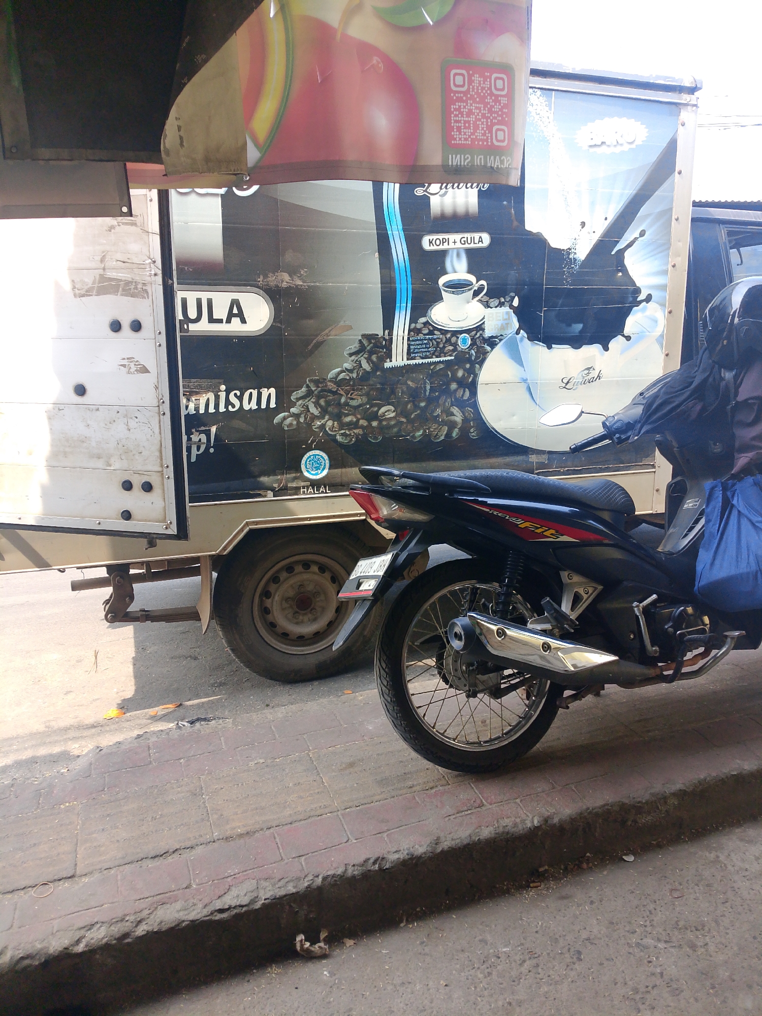 A parked motorcycle is visible in the foreground of the image, positioned on a sidewalk adjacent to a truck. The truck has a large advertisement on its side, featuring an image of coffee beans and a cup of coffee, along with text in Indonesian. The motorcycle is black with red and silver accents and has a blue bag hanging from its rear. The truck is white and appears to be a delivery vehicle. The scene is set in an urban environment, with a paved road and sidewalk. The presence of the truck and motorcycle suggests a commercial or transportation-related setting, possibly a market or a delivery route in Palembang, Indonesia. The sun is shining, casting shadows on the ground.