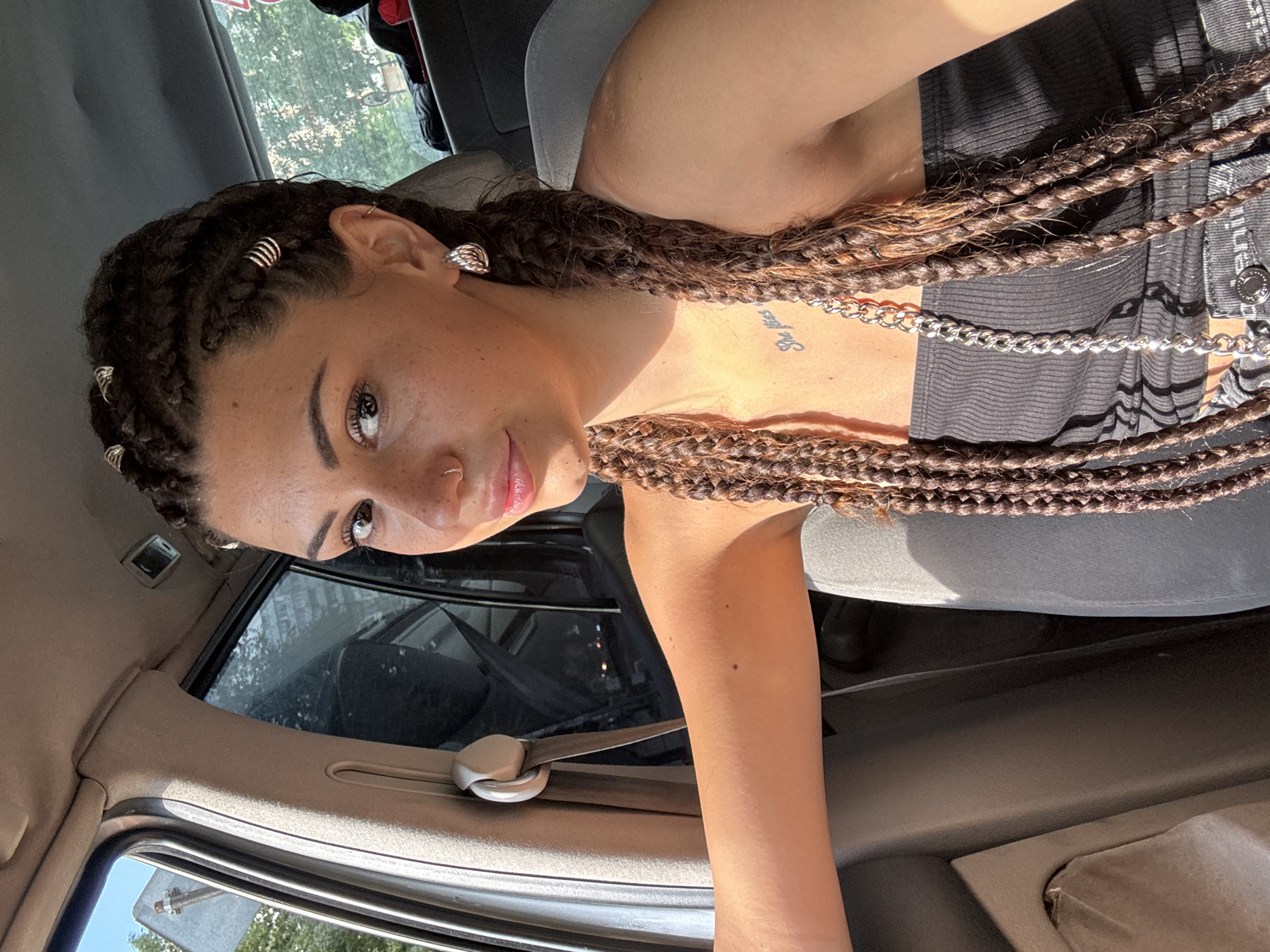 A young woman with braided hair, adorned with silver rings, sits in the driver's seat of a car. She is wearing a black tank top and a silver chain necklace, with her arm resting on the steering wheel. Sunlight streams through the car window, illuminating her face and freckles. The interior of the car appears to be fabric-covered and practical. Outside the window, trees and buildings are visible, suggesting an urban setting. The woman is looking at the camera with a slight smile. The image is a selfie taken inside the car.