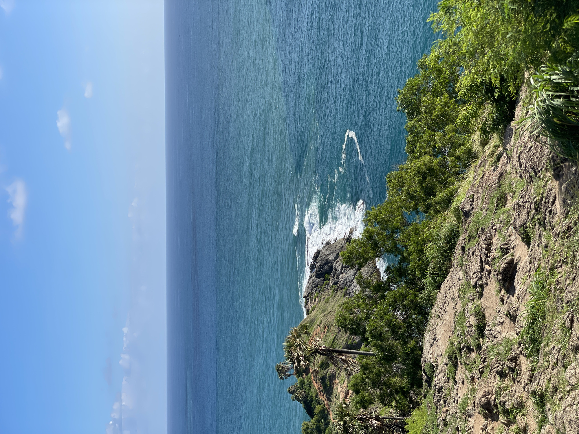 The image depicts a panoramic view of the ocean from a rocky, vegetated cliffside in Rawai, Thailand. The foreground is dominated by rugged, dry terrain sparsely covered with green grass and small plants. Further back, lush green trees and several palm trees with their distinctive fronds are visible, clinging to the slopes of the headland. In the middle ground, a rocky outcrop juts into the sea, where waves crash against the shore, creating white foam and spray. The turquoise blue ocean stretches out to the horizon, meeting a clear blue sky dotted with a few white clouds. The overall scene conveys a sense of natural coastal beauty.