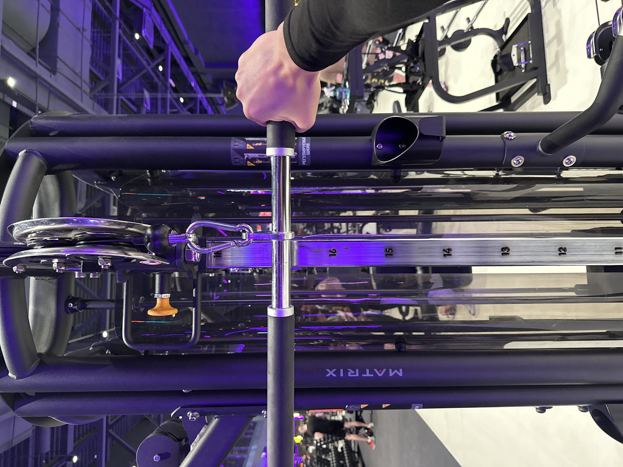 A person's hand grips a black-handled metal pin, which is part of a "Matrix" branded exercise machine. The machine has a pulley system with numbered height adjustments. The background shows elements of a modern interior with blue and white lighting. There is no indication that the image was taken in Murino, Russia.