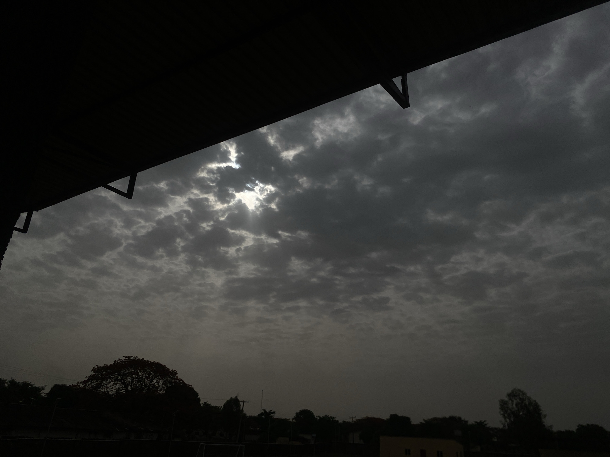 The image depicts a cloudy, overcast sky in Tudun Wada, Nigeria. The sky is filled with dark, mottled clouds, with a small break allowing some sunlight to peek through. In the foreground, the edge of a dark roof is visible, obscuring the view. Below the sky, a line of trees and buildings is silhouetted against the cloudy backdrop. A football field with goalposts is faintly visible, near a house. The overall lighting is dim, indicative of a stormy or late-day scene. There are no visible people or immediate actions taking place in the frame.