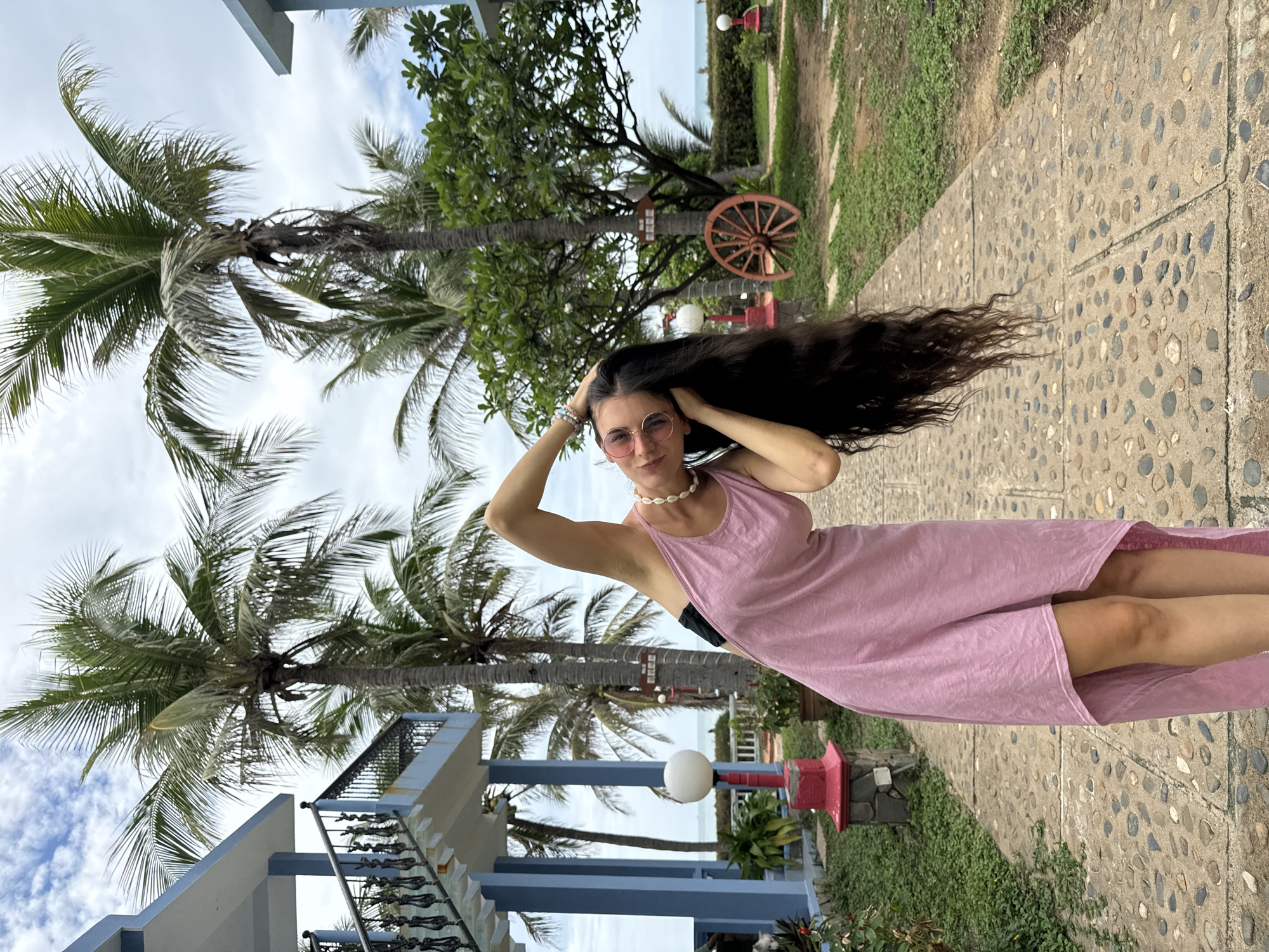 A young woman stands outdoors in a tropical setting, posing for a photograph. The image is oriented vertically, with her body angled slightly towards the camera and her head turned to face it directly. She has long, dark, wavy hair that cascades over her left shoulder, and her hands are raised to her head, as if she is running her fingers through her hair. She wears a light pink sleeveless dress, a white shell necklace, and round, light-colored sunglasses. A subtle smile plays on her lips, conveying a relaxed and confident demeanor.
The background features lush tropical vegetation, dominated by numerous tall palm trees with green fronds. To the left, parts of a building with blue and white trim and a dark-railed staircase are visible. In the mid-ground, a paved walkway made of embedded stones stretches across the lower portion of the image, flanked by green grass and bushes. Decorative elements include white spherical lamp posts, one with a red base, and a rustic wooden wagon wheel mounted upright against a tree trunk. In the distance, beyond the trees and structures, a glimpse of the ocean can be seen, indicating a coastal location.
The sky is bright but mostly overcast with visible clouds, suggesting daytime with soft, diffused light, possibly late morning or afternoon. The overall scene evokes a resort or coastal garden environment, consistent with the provided location context of Mũi Né, Vietnam. No legible text is visible on any signs or objects.