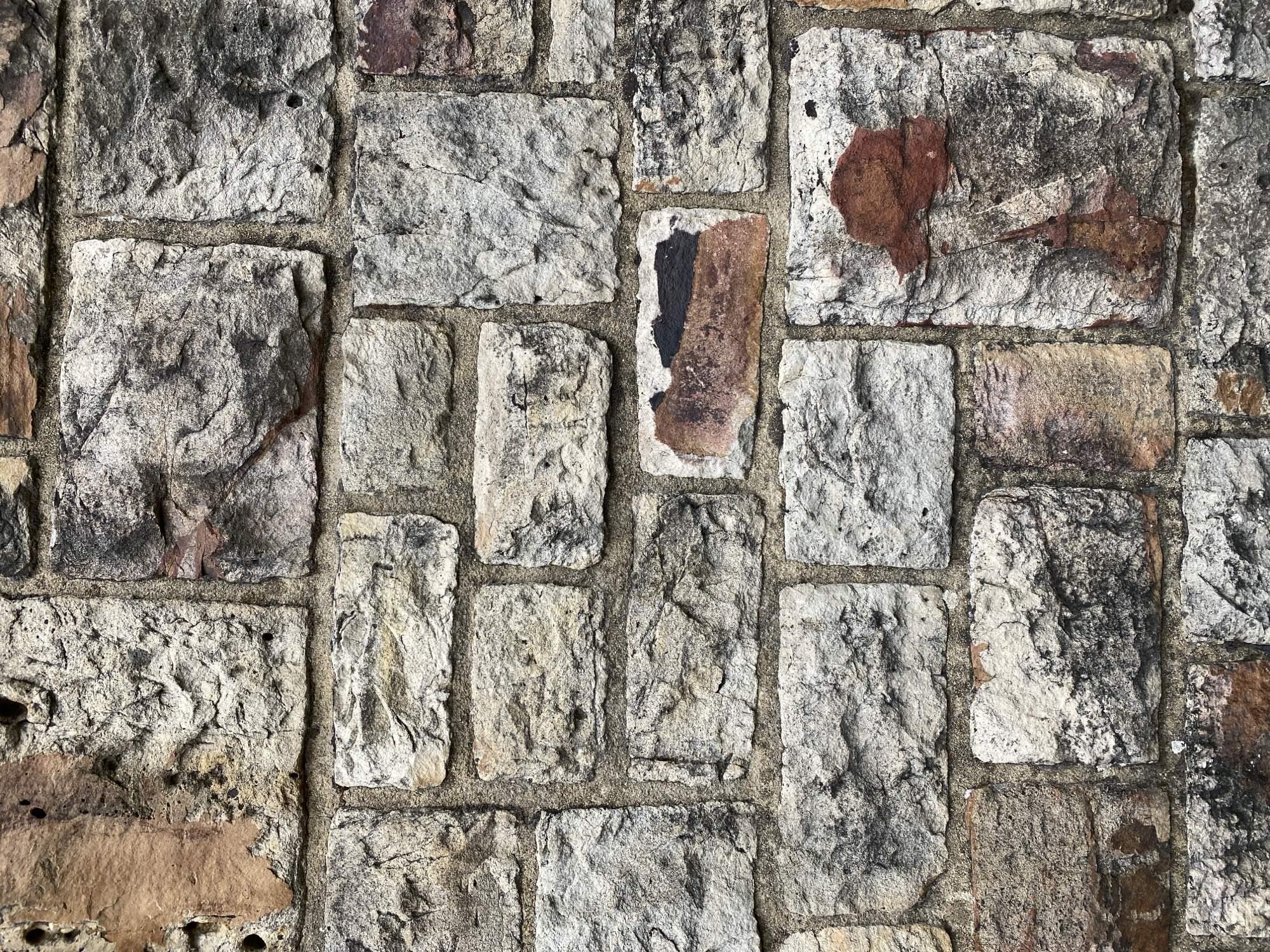 The image is a close-up, head-on, full-frame photograph of a weathered stone wall. The stones are of varying sizes and irregular shapes, laid in a coursed rubble pattern with a light-colored mortar filling the gaps between them. The stones exhibit a range of colors, including shades of gray, beige, white, and reddish-brown, with textures that are rough, pitted, and chipped. Some stones have darker grey or blackish patches, possibly from lichen or staining. There are small, dark circular holes visible in some of the stones. The overall impression is of an old, rustic, and durable structure. The photograph does not contain any people, animals, or discernable landmarks to pinpoint a specific location within Altenkirchen, Germany.