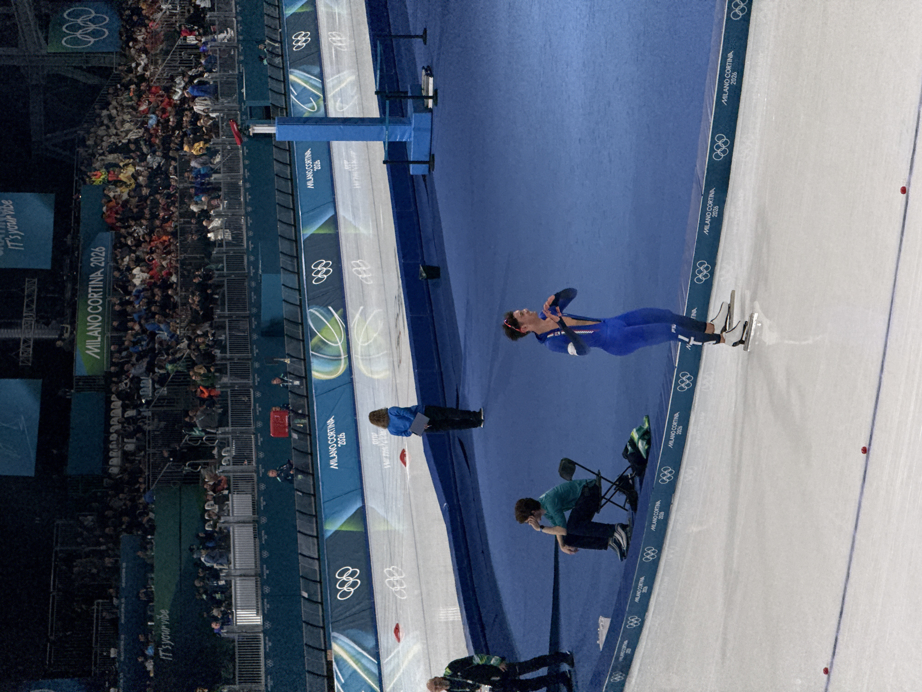 The image captures an indoor ice rink scene, likely an Olympic event. A speed skater in a blue suit is mid-motion on the ice, with Olympic rings and "MILANO CORTINA 2026" repeated along the rink's edge. Spectators fill the stands in the background. Officials and personnel are present near the rink's side, one seated and another with a clipboard. The rink's design incorporates blue and white colors. The phrase "It's your vibe" appears in the background.