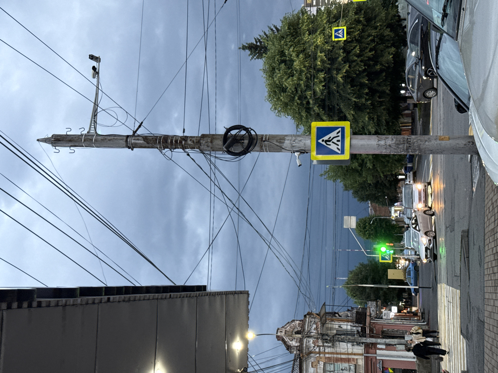 In the city of Assinovskaya, Russia, a street scene unfolds under a cloudy sky. A pedestrian crossing is visible on the left, with several individuals crossing. Cars are present on the road, some of which are moving, indicating traffic flow. A traffic light shows a green signal, suggesting that pedestrians currently have the right of way or that vehicles can proceed. A utility pole stands prominently in the foreground, supporting numerous wires and a surveillance camera. Buildings line the street, displaying architectural details consistent with urban environments. Lush green trees are interspersed throughout the scene, adding a natural element to the cityscape. The overall atmosphere is one of everyday urban activity, possibly during the late afternoon or early evening, given the lighting conditions.